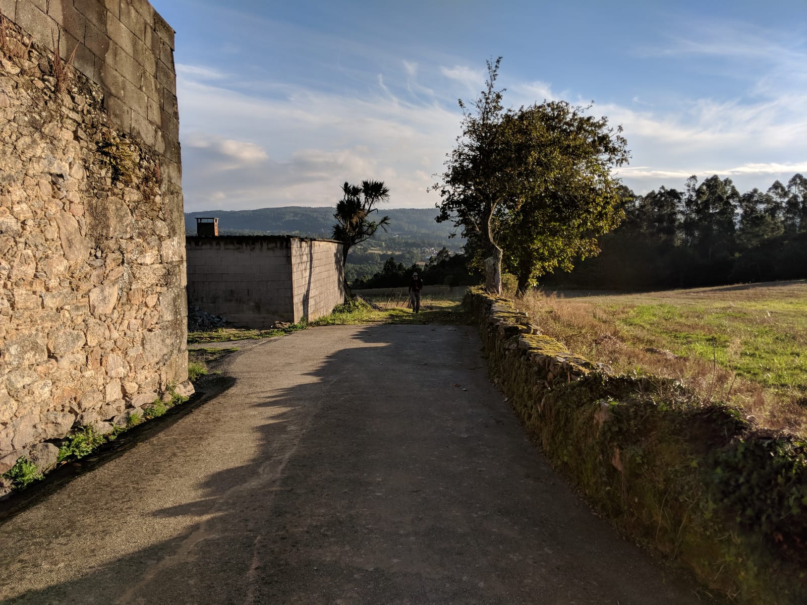 Paved lane past a stone building with countryside views and a pilgrim ahead