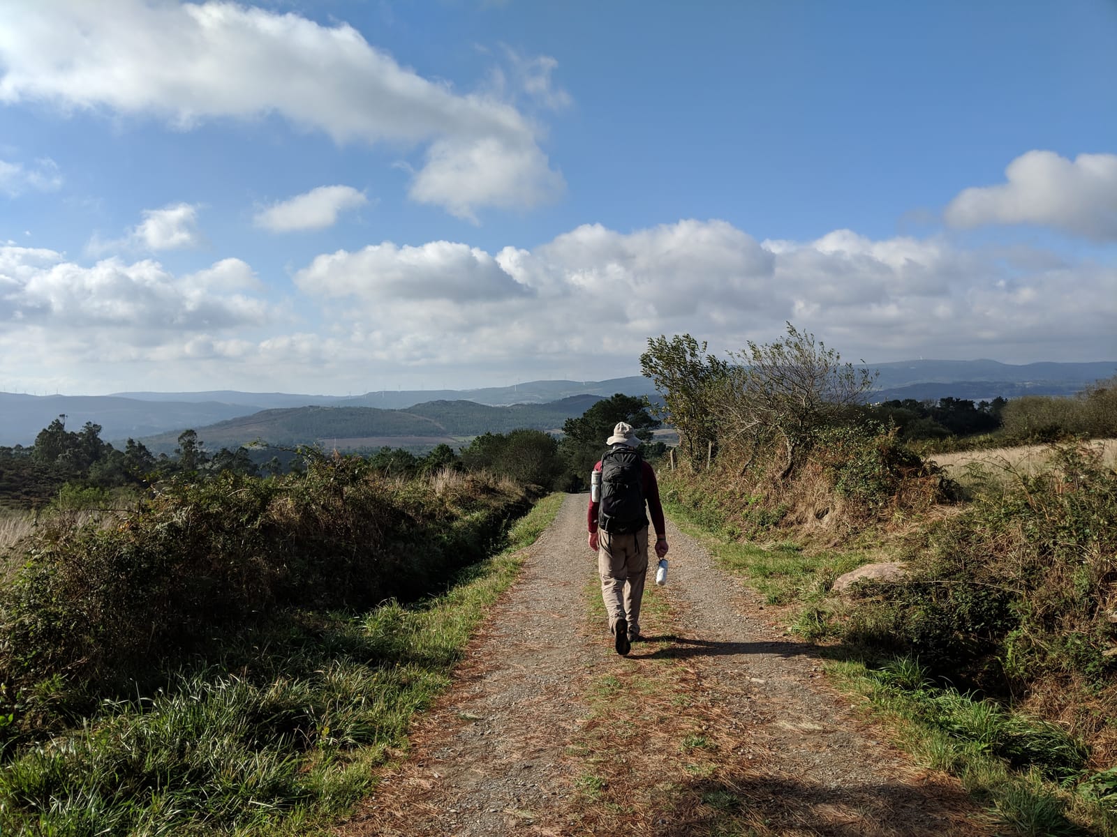Doug walking a gravel ridge path with sweeping views of distant hills and wind turbines
