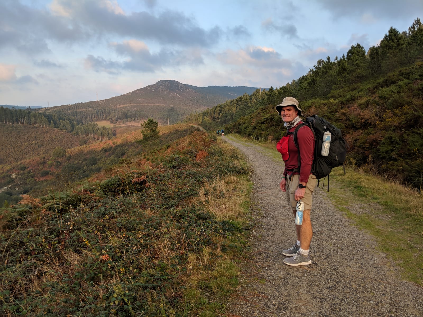 Mike pausing on a gravel ridge path with a peaked hill and pilgrims in the distance