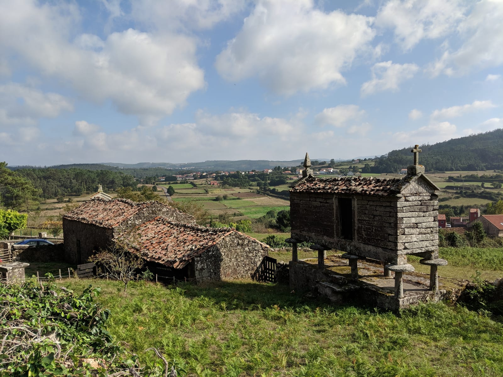 A granite hórreo with a cross beside an old stone house overlooking a green valley