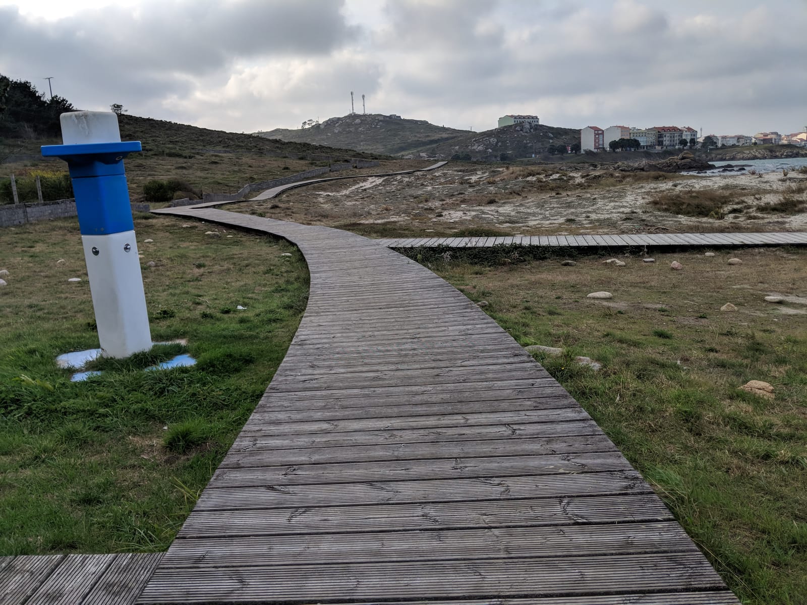 A wooden boardwalk crossing coastal dunes toward the town of Muxía under a gray sky
