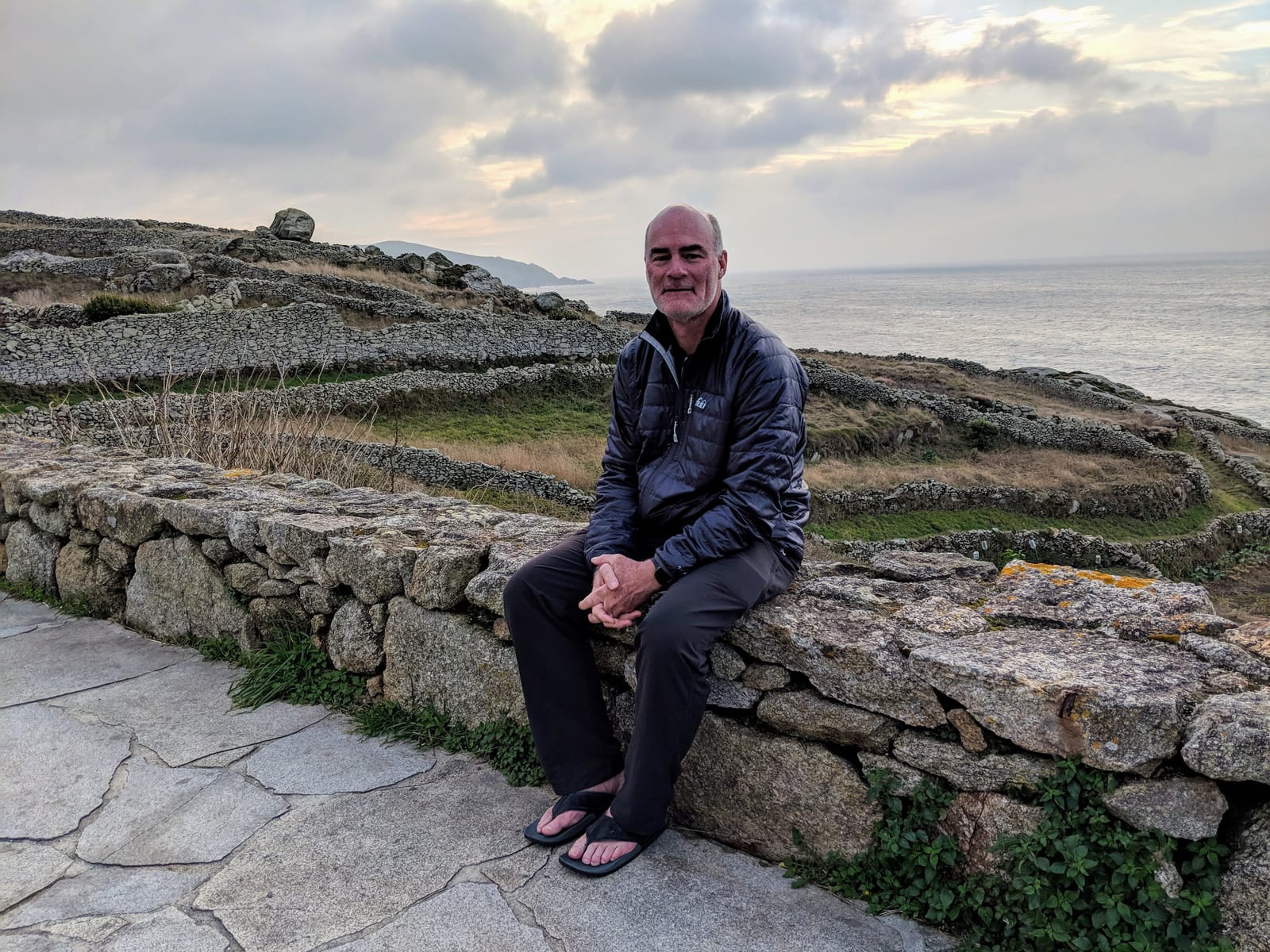 Doug on a stone wall at the Muxía point with the sea behind