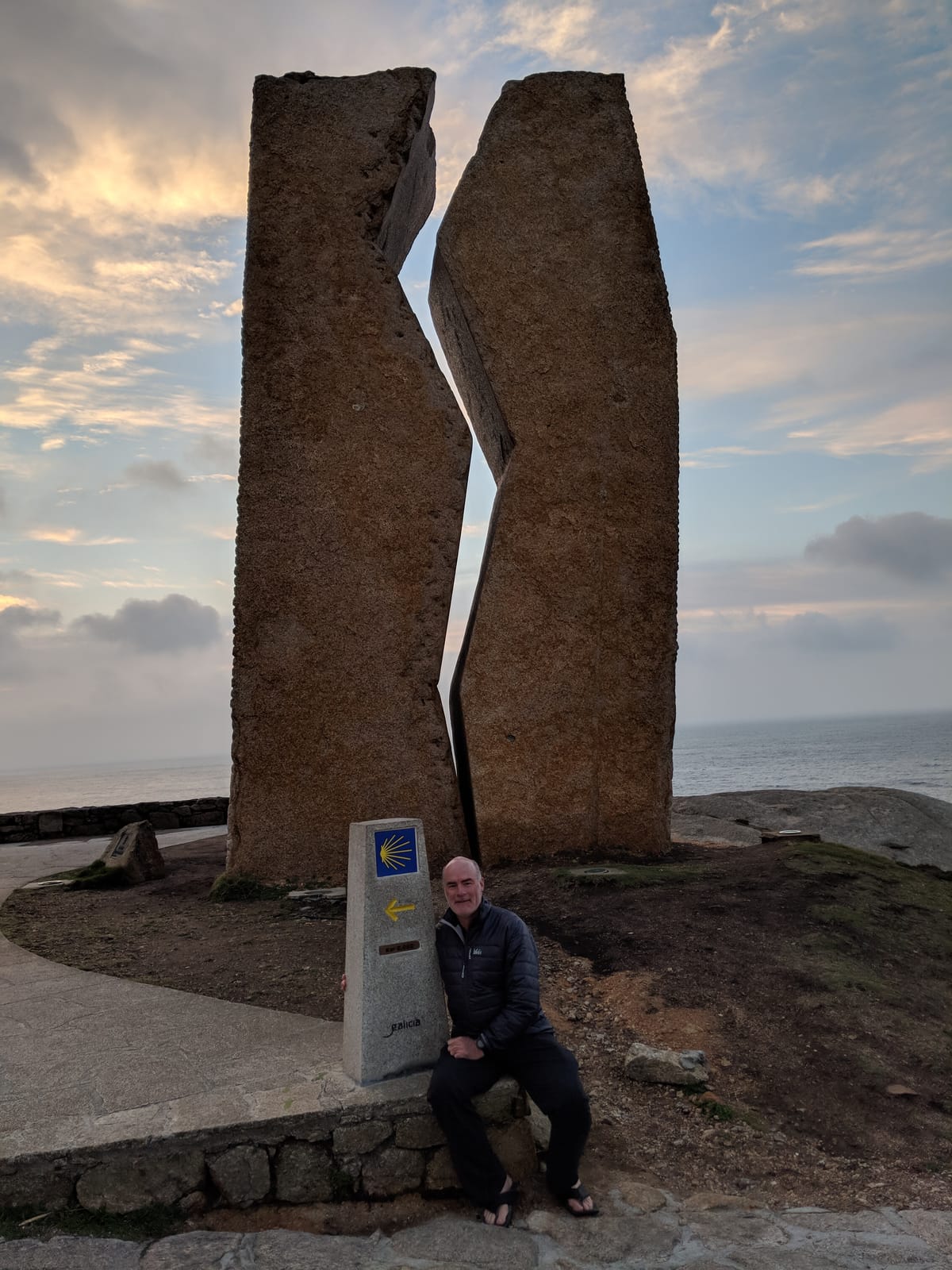 The tall split-granite sculpture A Ferida on the Muxía coast, with a Camino marker at its base