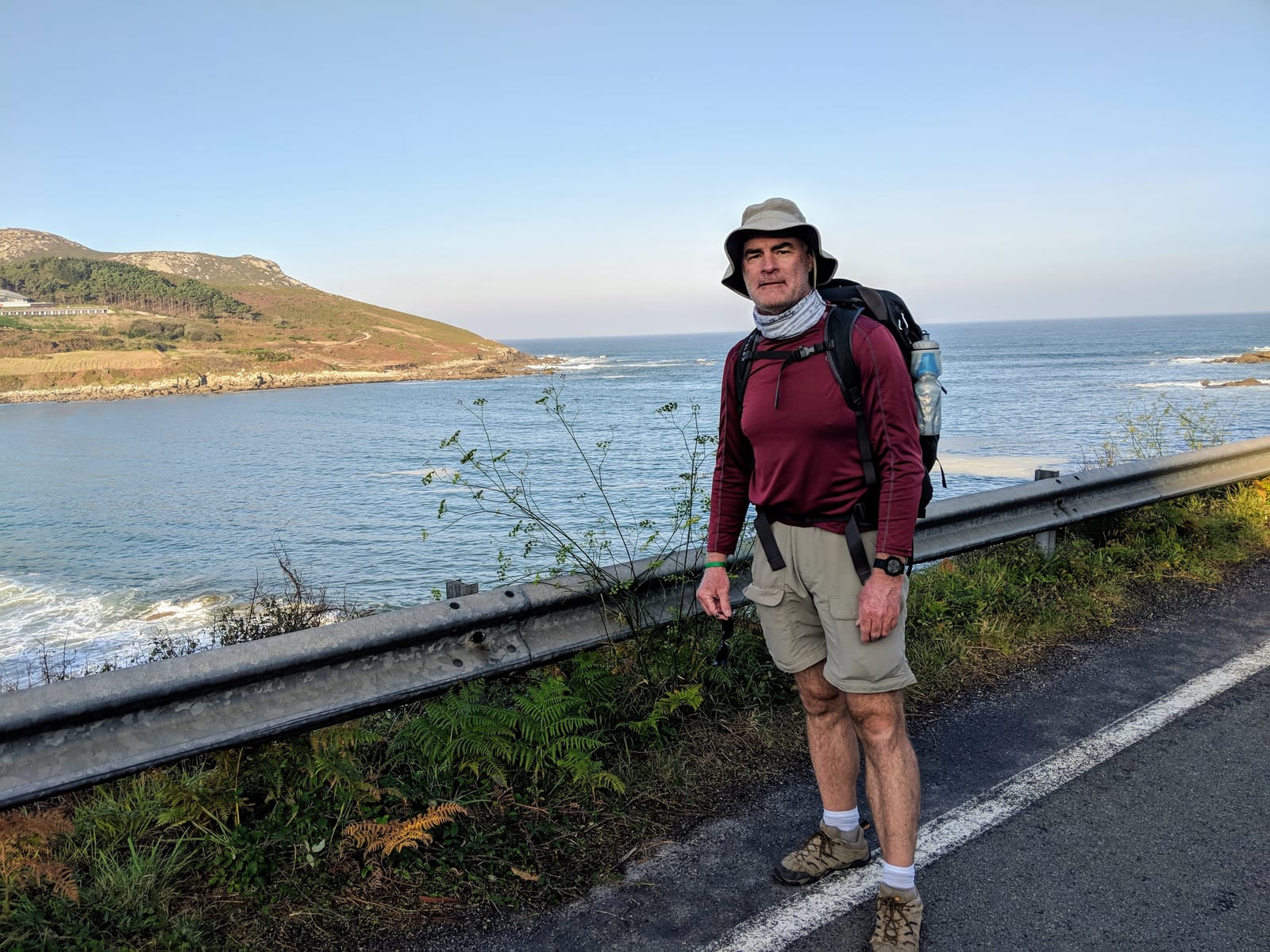 Doug pausing at a guardrail on the coast road above a bay, a green headland behind him