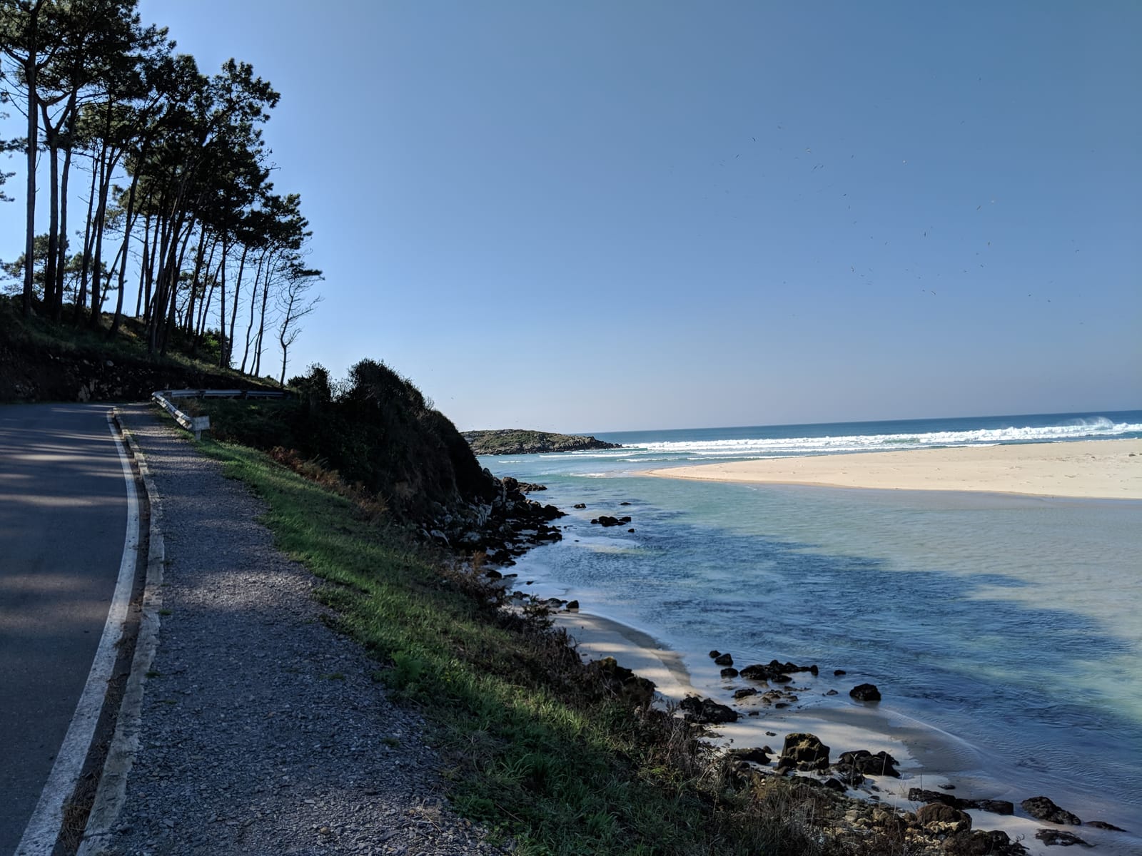 A coast road curving above a turquoise bay with a long pale-sand beach and breaking surf