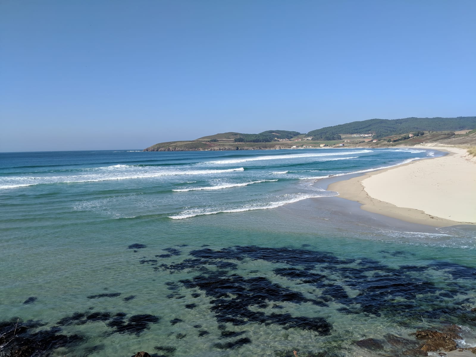 Clear turquoise water over dark rocks and kelp along a wide Galician beach