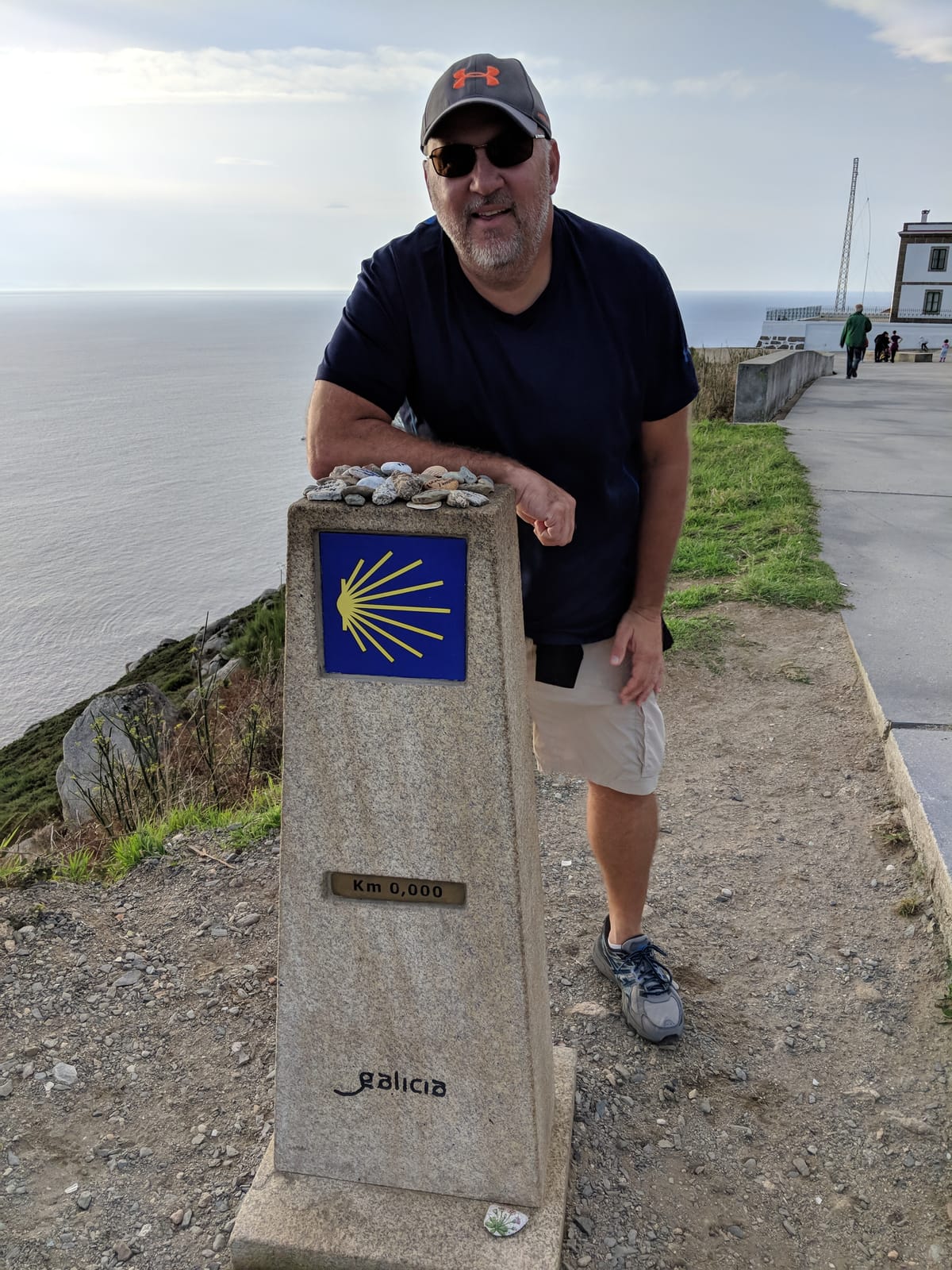 Mike leaning on the Km 0,000 marker with the Atlantic and cliffs behind
