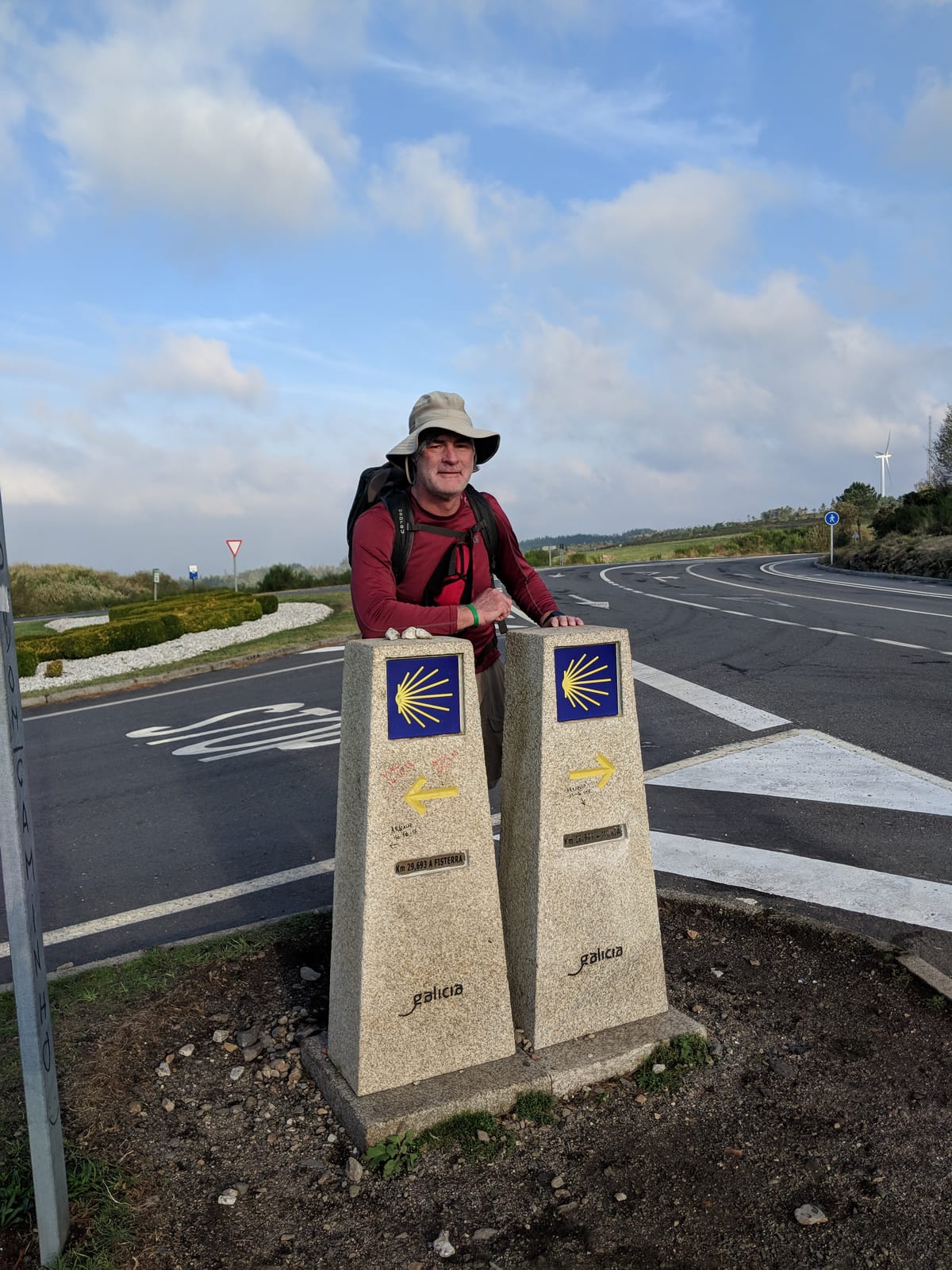 Mike leaning between two Camino waymarker pillars, one arrow pointing left to Fisterra and one right to Muxía