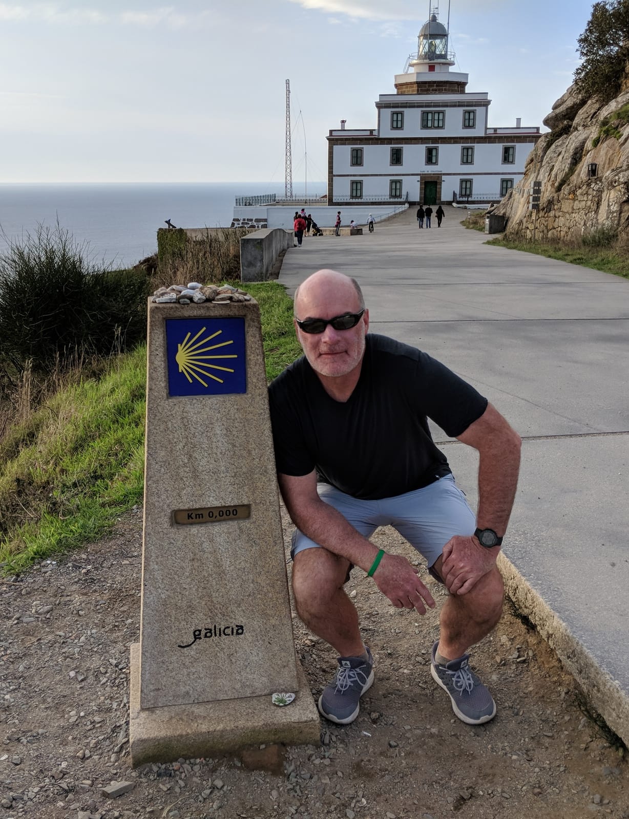 Doug crouched beside the Km 0,000 marker with the Finisterre lighthouse behind