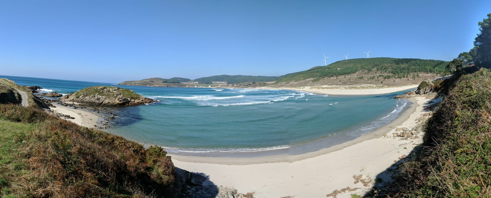 Panorama of a curving turquoise cove with pale sand, rocky islets and wind turbines on the hills