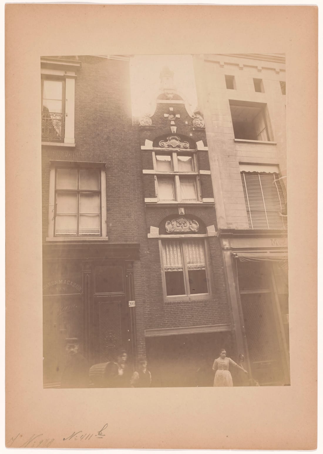 Faded mounted photograph of the same corner, showing on the left a plain brick front with tall sash windows, a shop window lettered with a word ending in -arenmagazijn van, and a panelled door with the number plate 209; in the centre the narrow gabled house; people in the street in the foreground