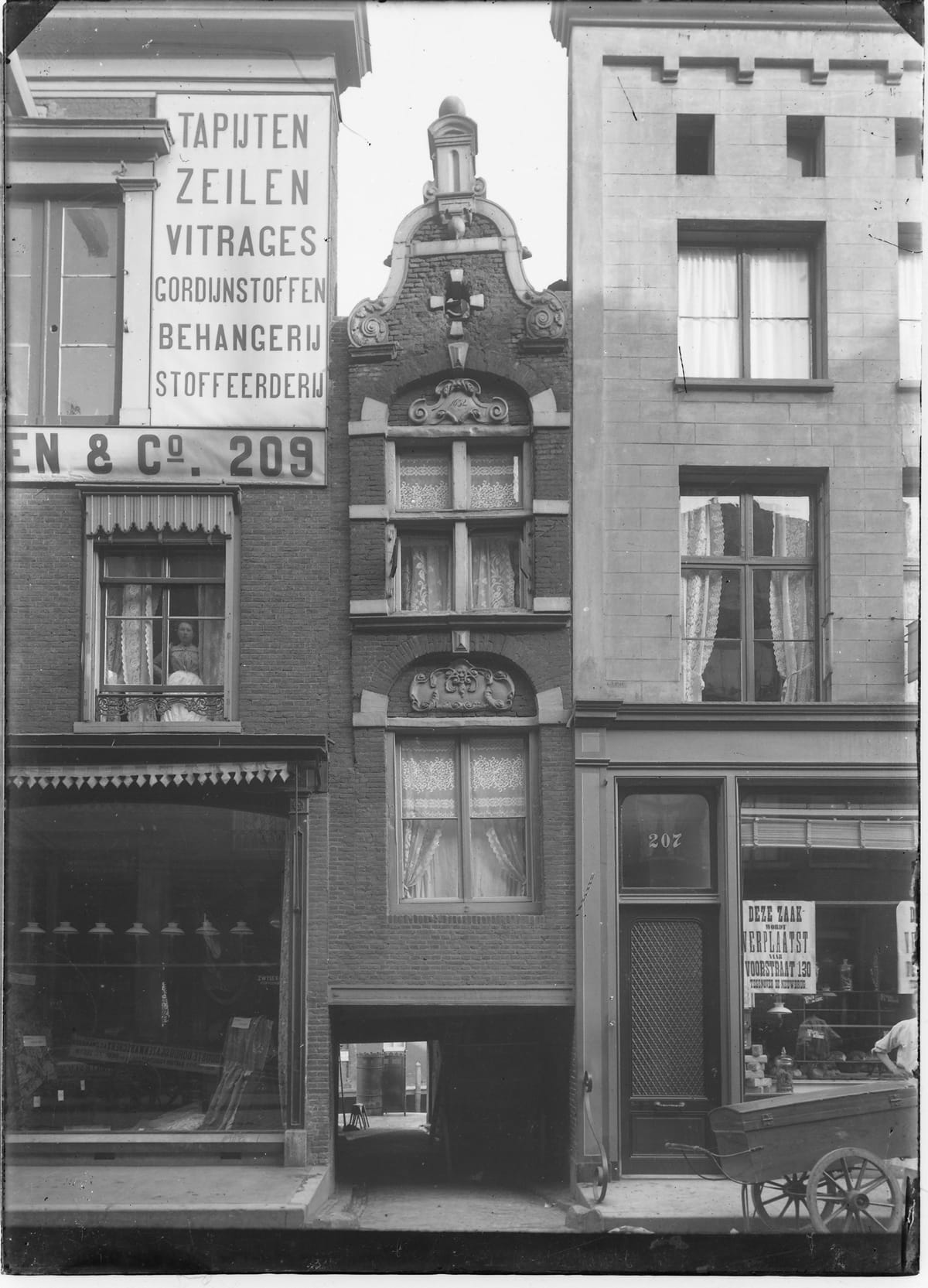 Glass-plate photograph of the narrow gabled house of 1632 over the dark alley entrance, flanked on the left by a brick shop-house with a big painted sign reading Tapijten, Zeilen, Vitrages, Gordijnstoffen, Behangerij, Stoffeerderij, En and Co. 209, a woman at its upstairs window, and on the right a door numbered 207