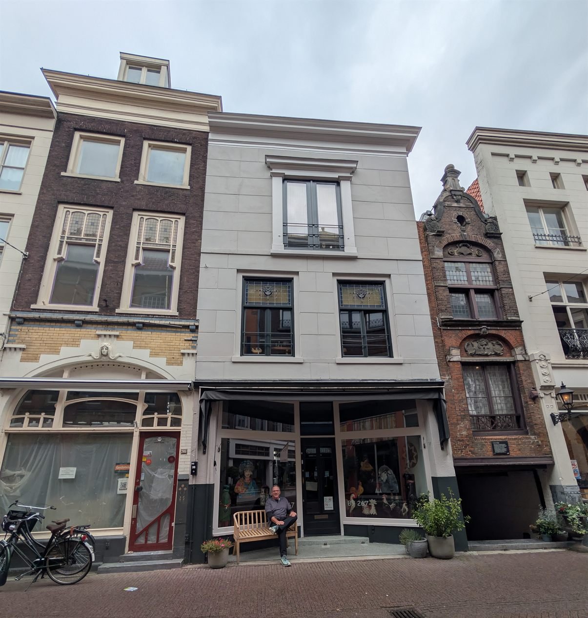The same house, with a man in a dark shirt sitting on the wooden bench outside the shop door