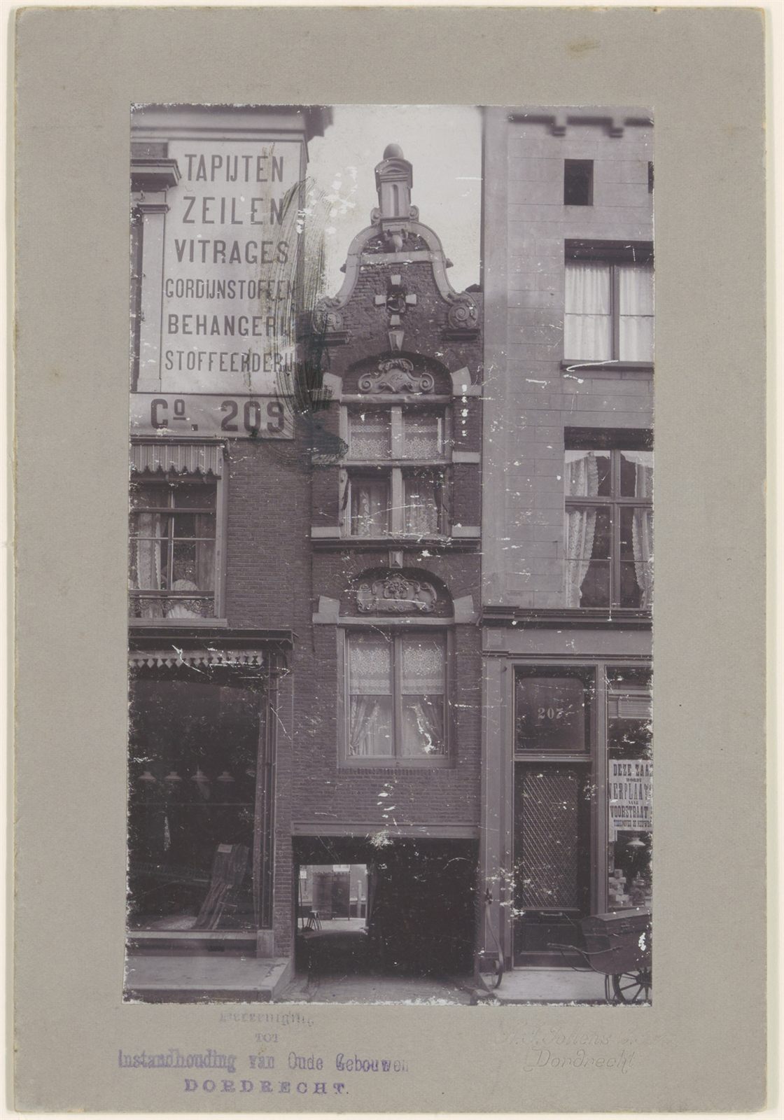 Old mounted photograph of the narrow gabled house over the alley, flanked by a shop wall painted with a list of trades ending in the number 209, and a door numbered 207