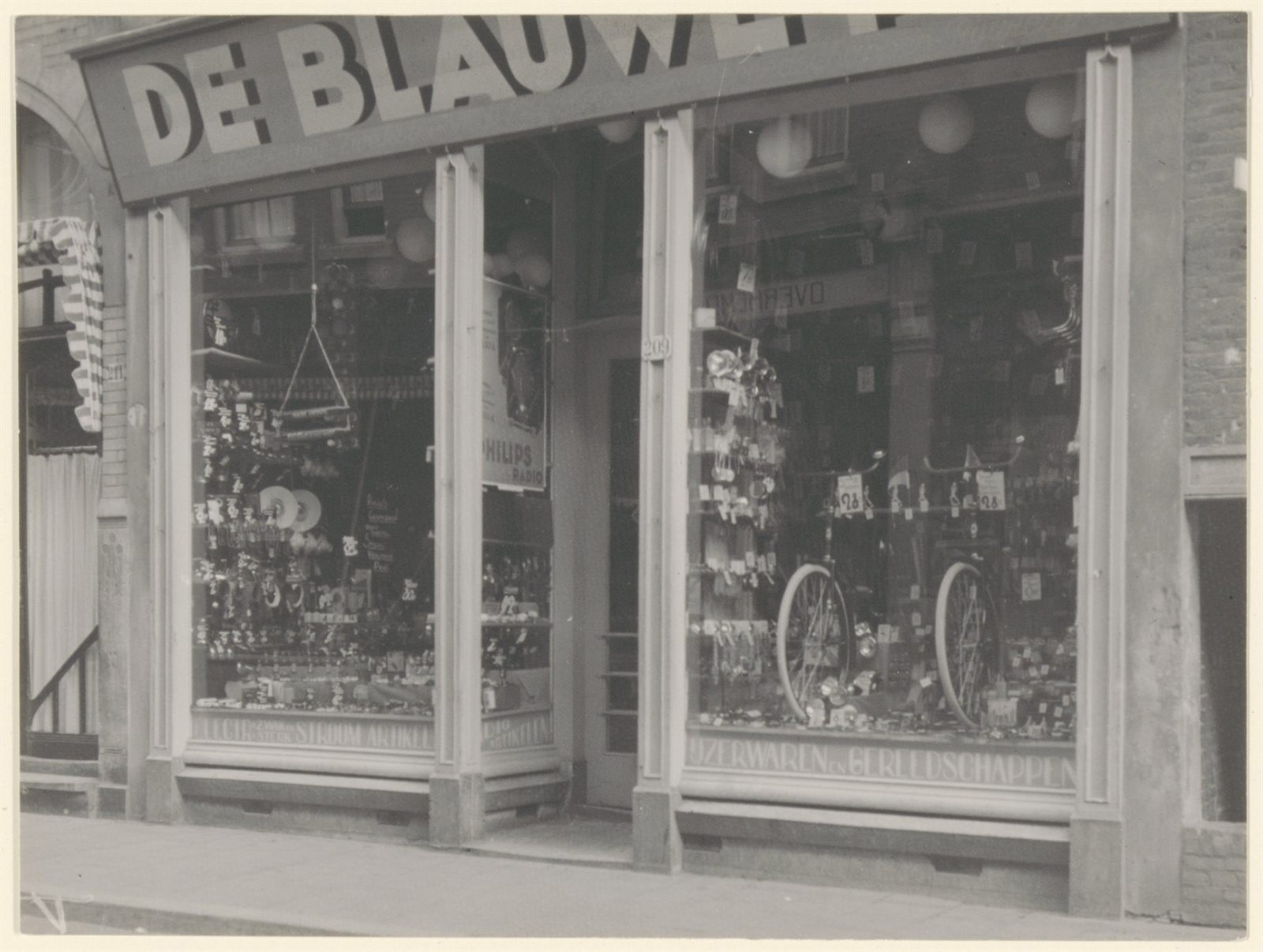 1930s photograph of an ironmonger's shopfront with two display windows flanking a central door, bicycles and hardware in the windows, and the number 209 beside the door