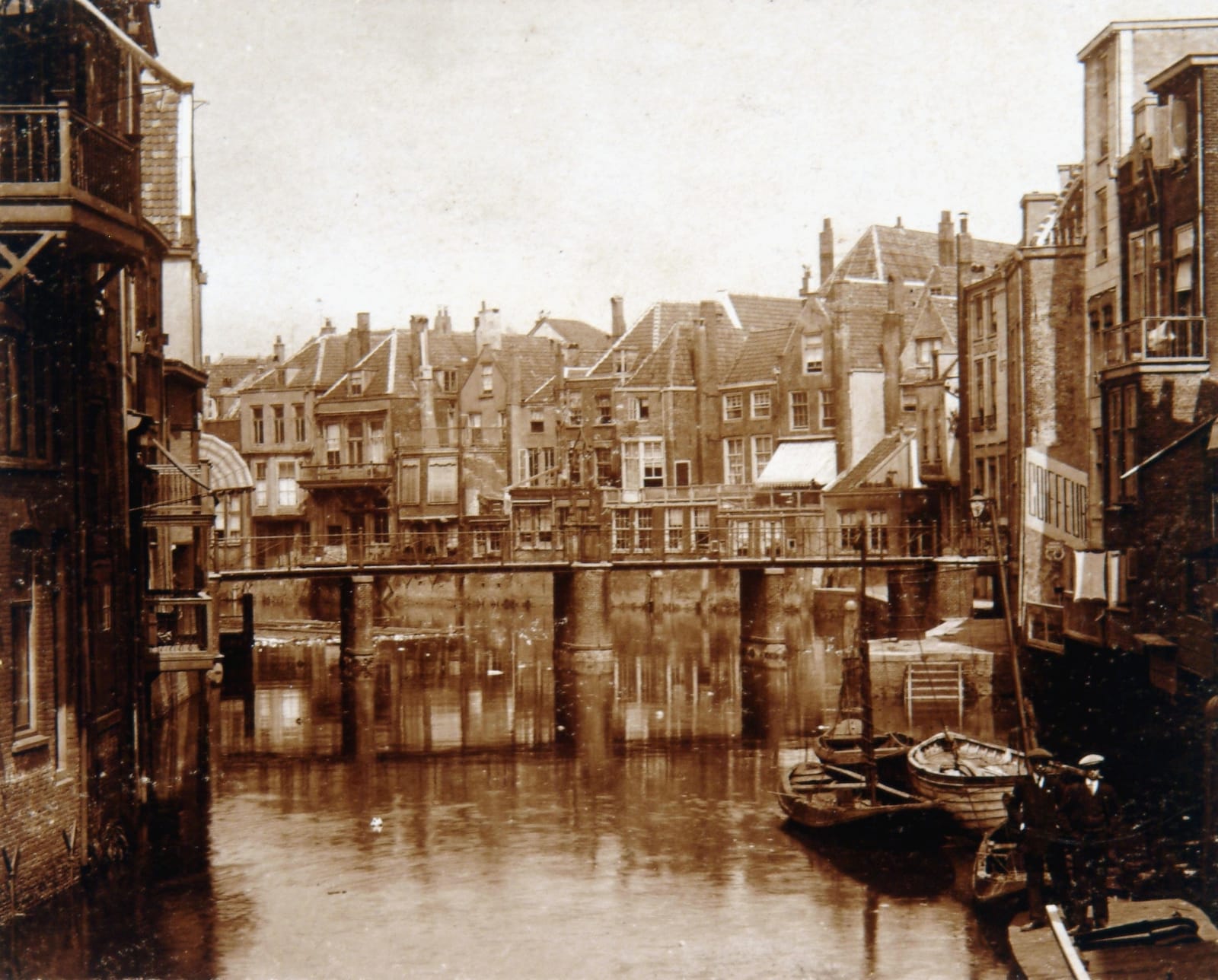 Sepia photograph of the Wijnhaven seen from the Tolbrug toward the Wijnbrug, a footbridge on piers crossing the water, rowing boats moored at the right, and on the right the backs of the Voorstraat houses, one with the word COIFFEUR painted down its side wall