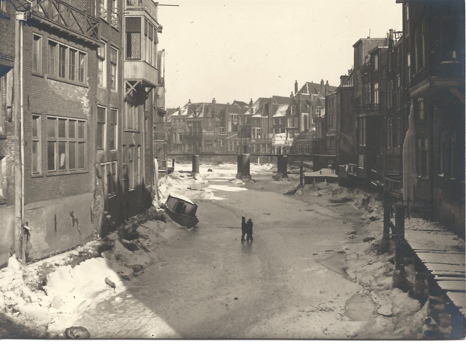 Old photograph of the Voorstraatshaven emptied of water in winter, snow on the banks, a boat lying over on the harbour bed and two figures standing in it