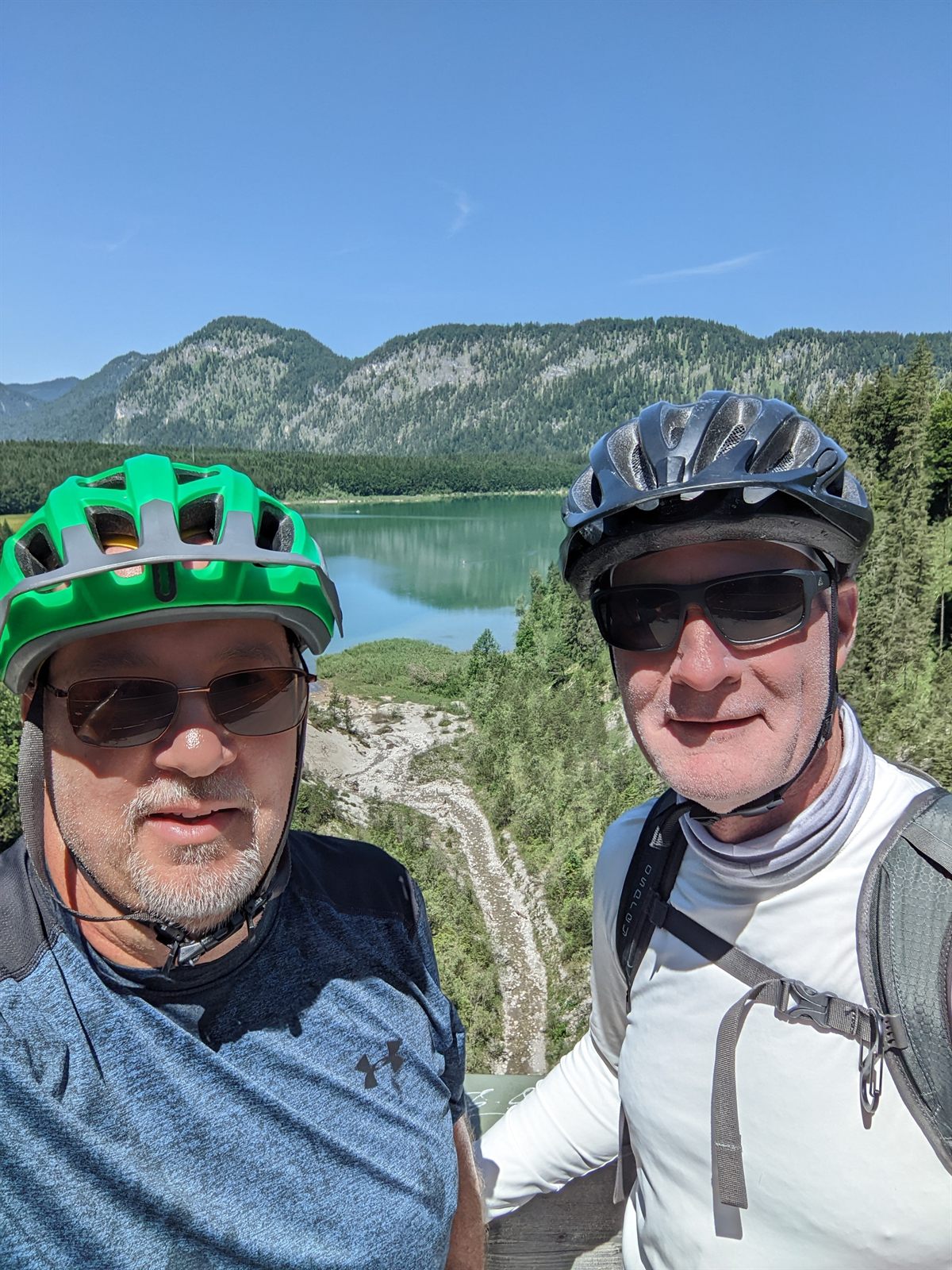 Selfie of Mike and Doug in cycling helmets above the turquoise Sylvenstein reservoir