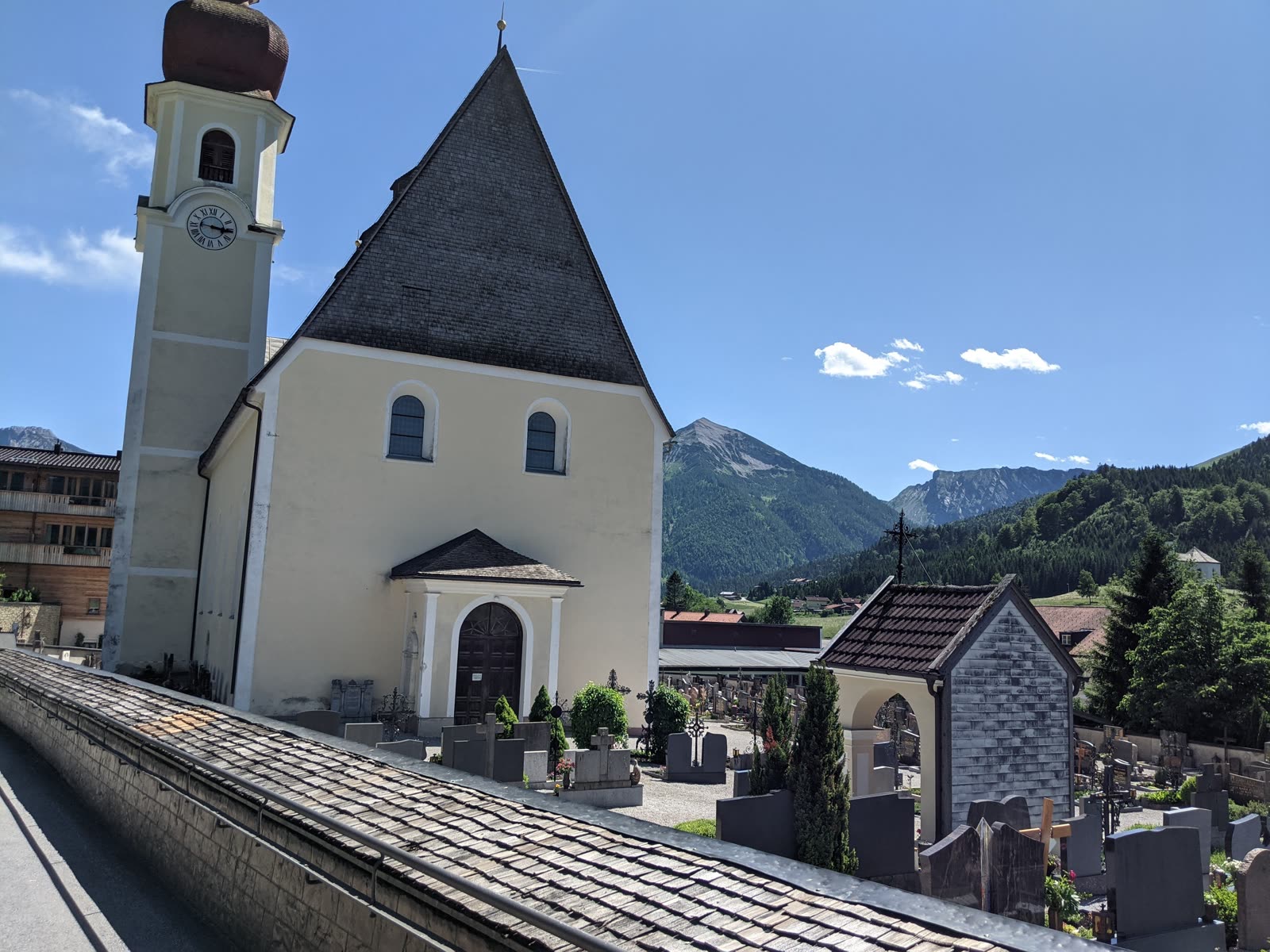 Tyrolean village church with an onion-dome bell tower and its hillside graveyard, a broad green valley and the Rofan peaks beyond