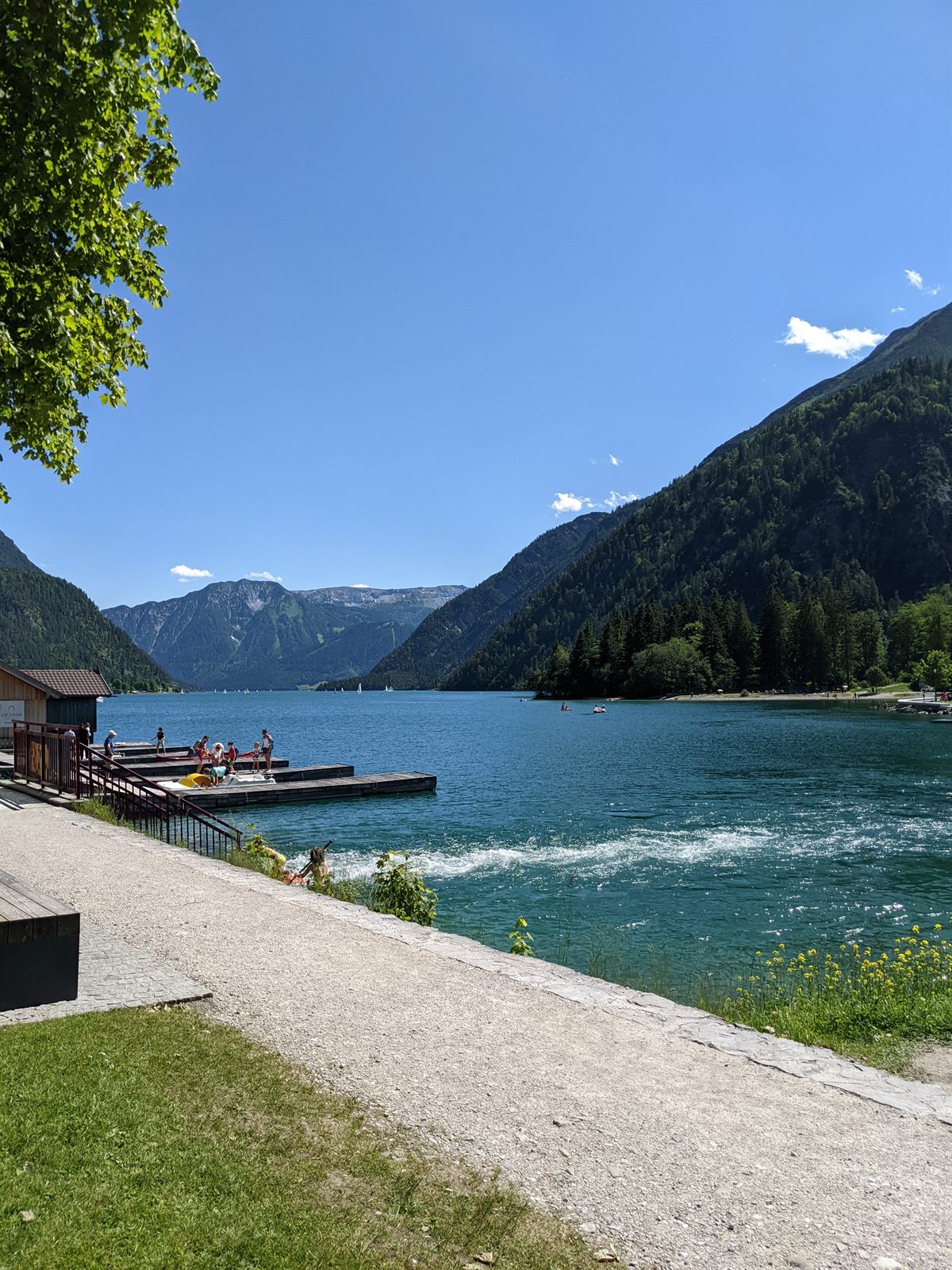 Wooden swimming jetty on the Achensee with people, sailboats beyond, mountains framing the lake