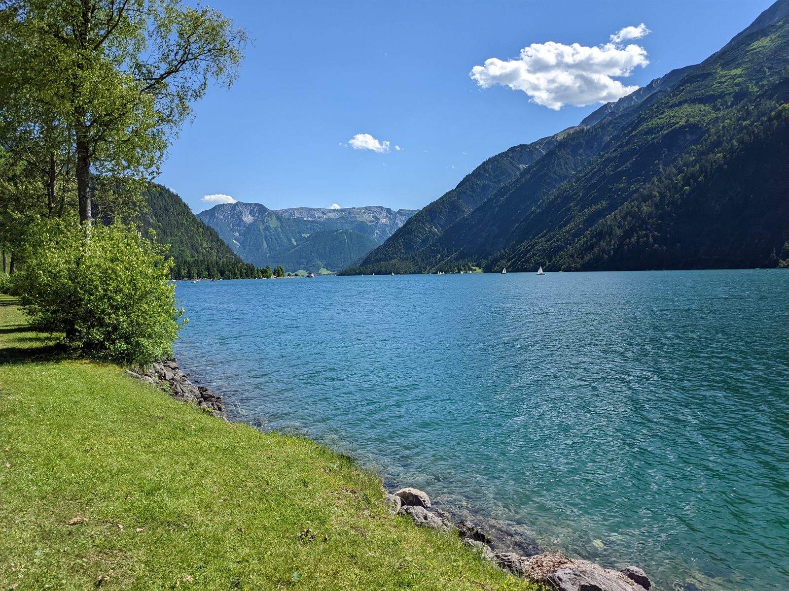 Wide view down the turquoise Achensee with grassy shore, sailboats, and steep forested mountains