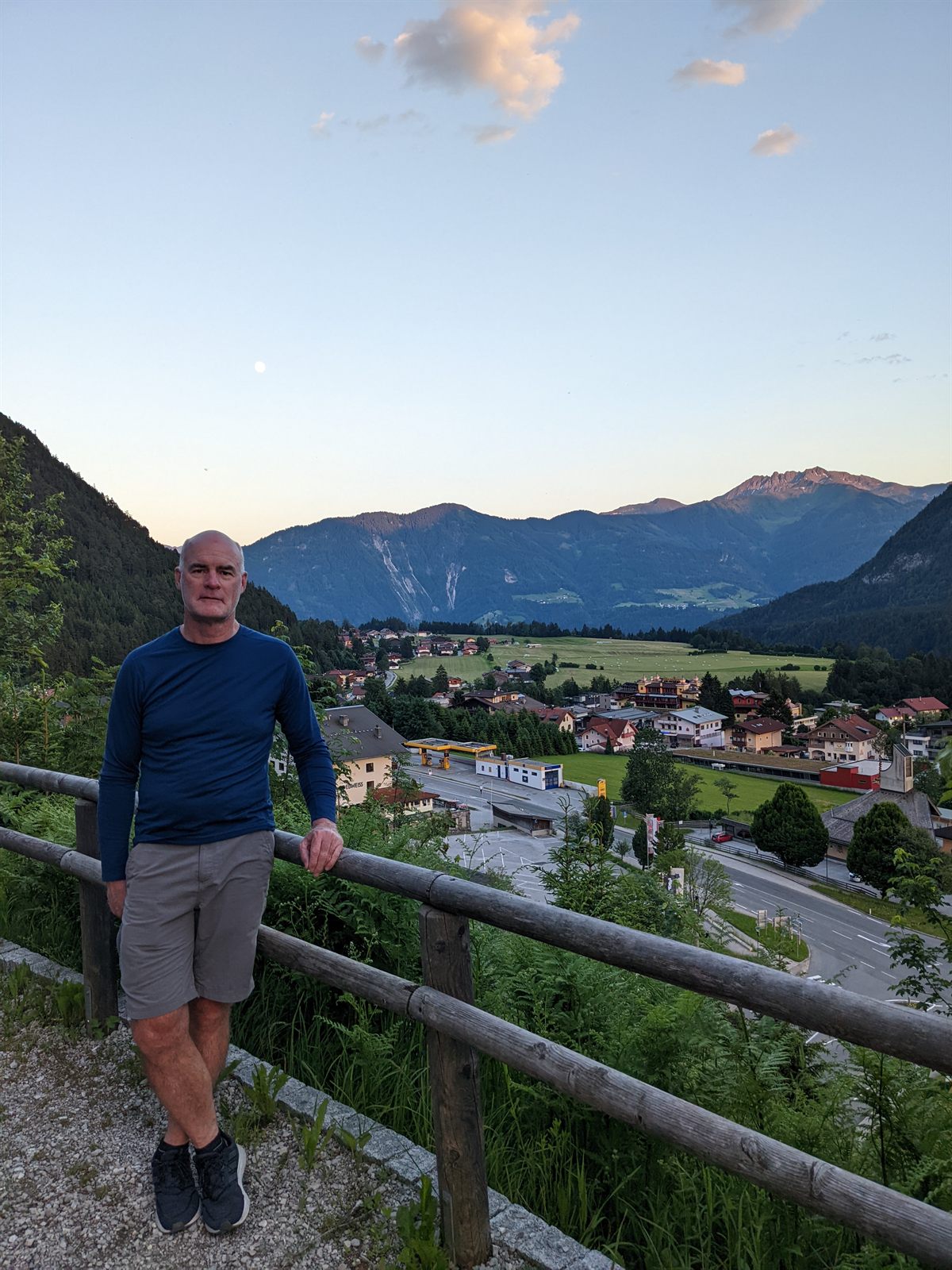 Doug leaning on a wooden railing above Maurach at dusk, a near-full moon rising over the valley