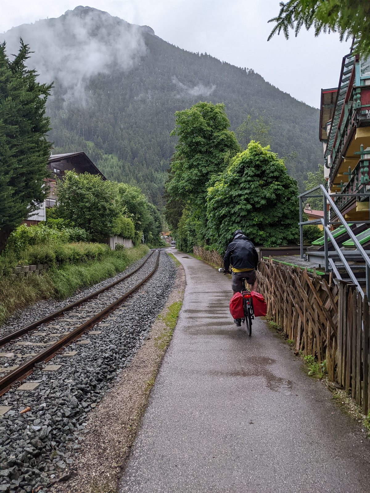 Doug in black rain gear riding a wet paved path beside old railway tracks, low cloud clinging to a forested mountain