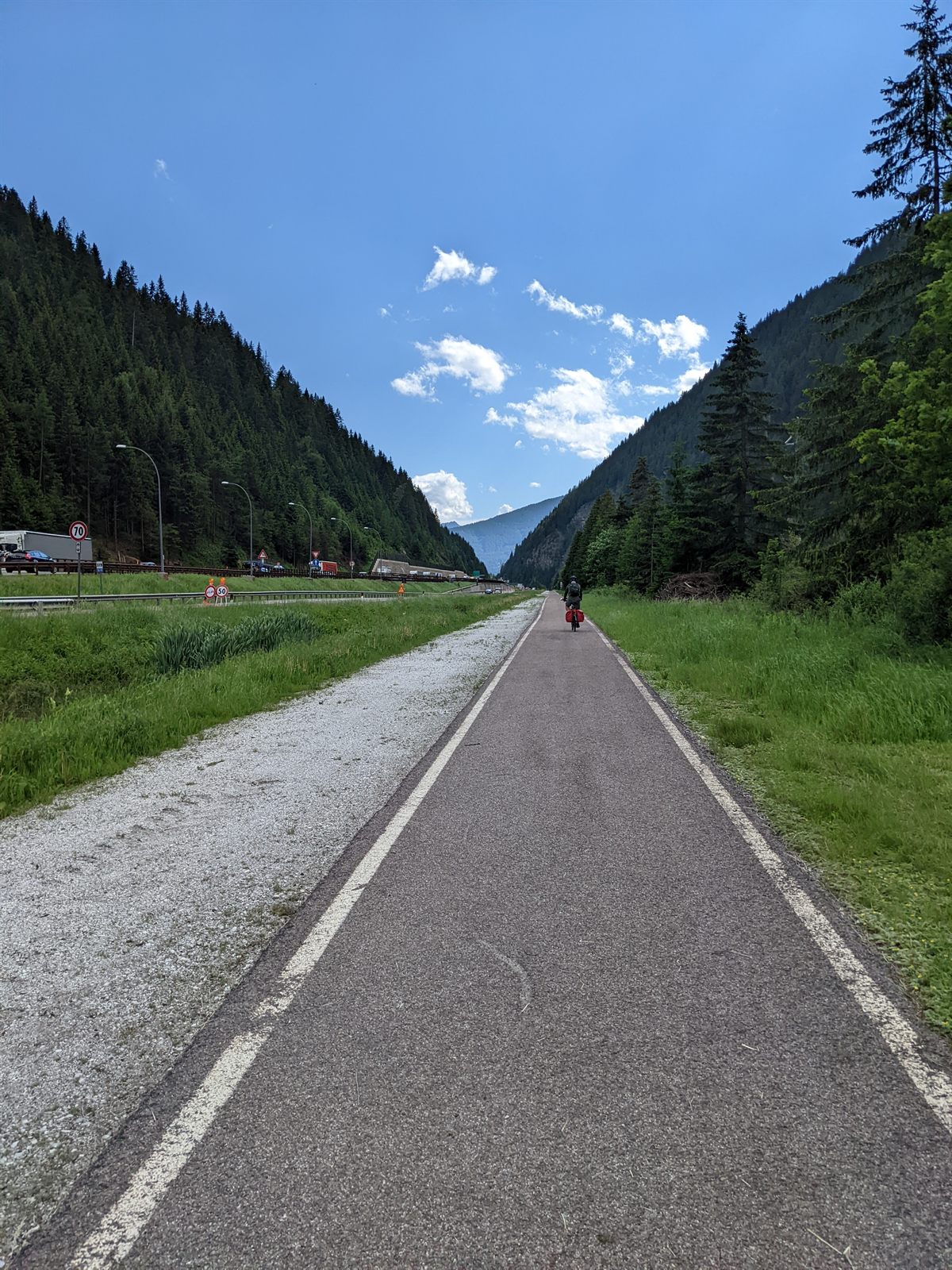Straight paved bike path running down a forested valley beside the autobahn, a loaded rider ahead under blue sky