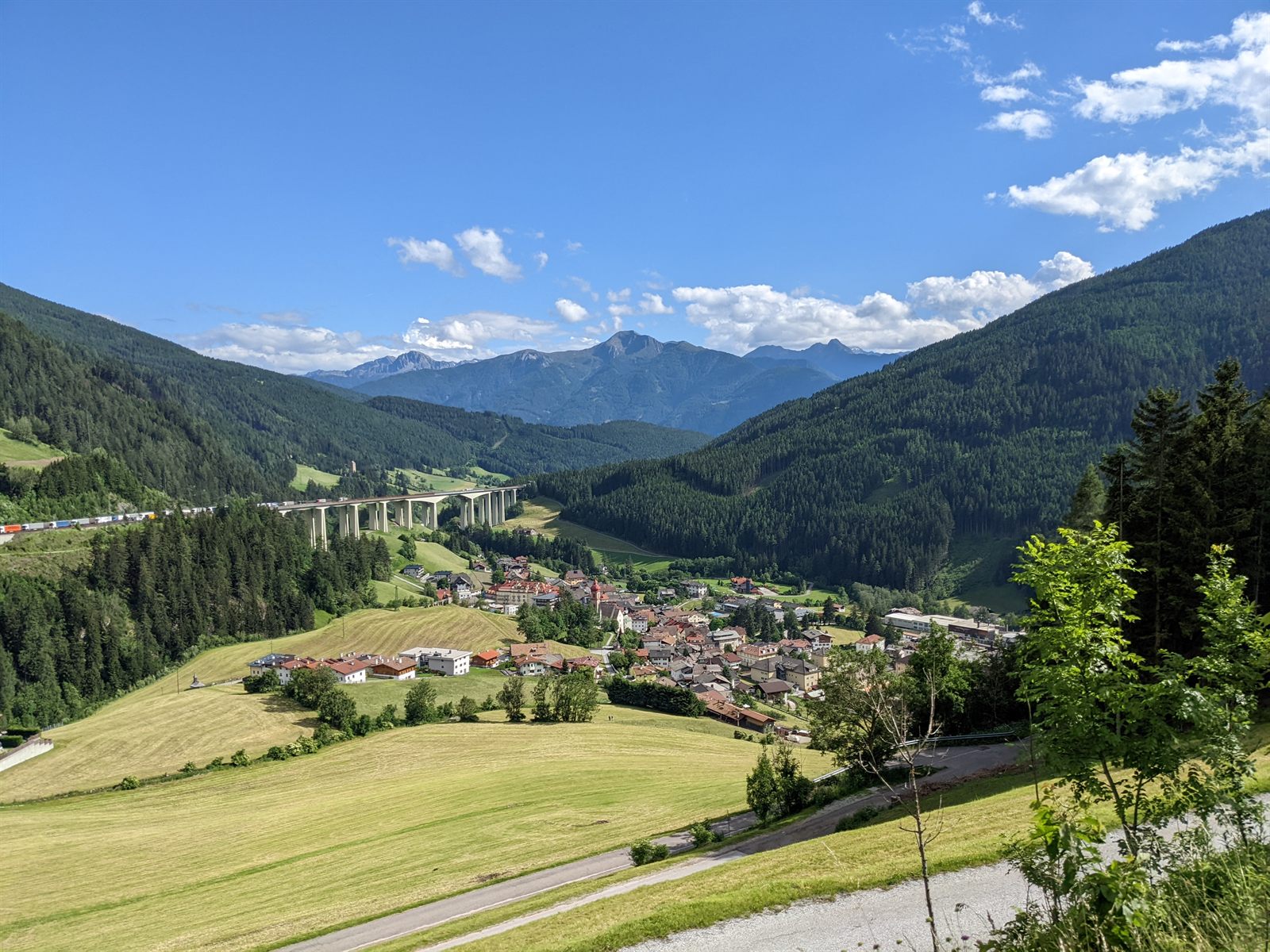 Wide view of a South Tyrol village in a green valley beneath the sweeping concrete viaducts of the Brenner autobahn, mountains behind