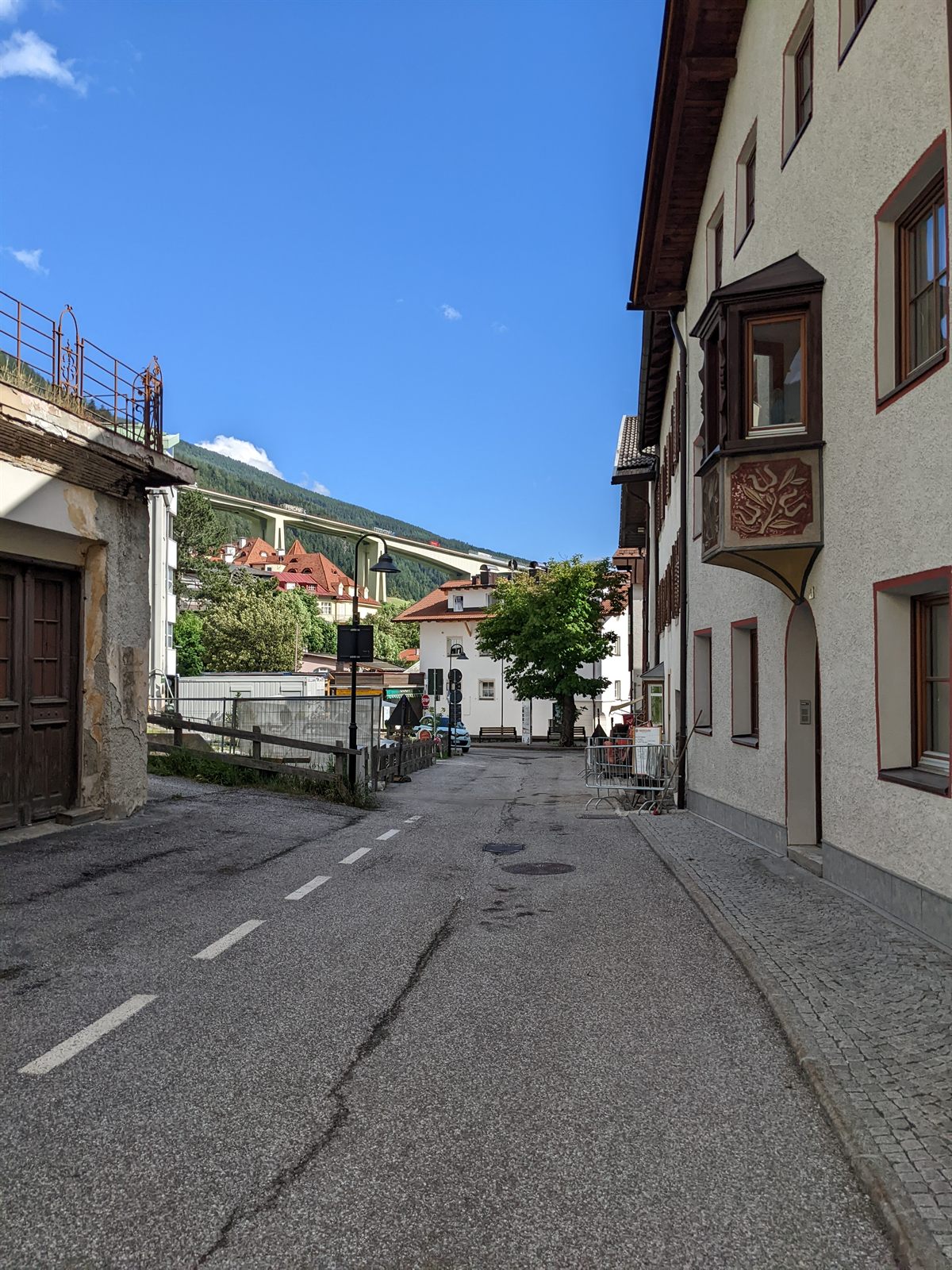 Quiet South Tyrol village street with a painted Tyrolean facade, the Brenner autobahn viaduct arcing across the hillside above