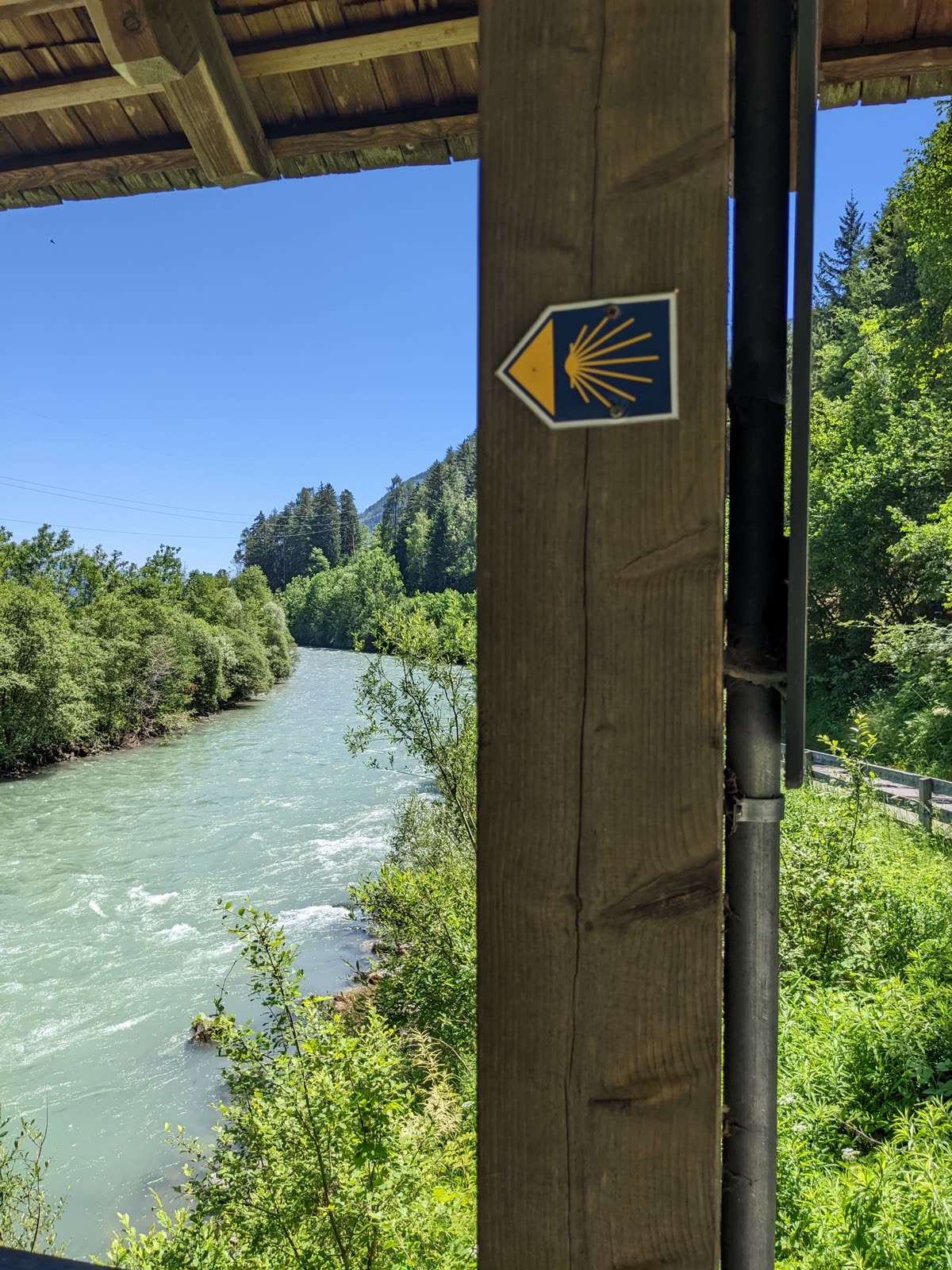 Yellow scallop-shell Camino waymarker on a blue plaque fixed to the post of a covered wooden bridge over a turquoise alpine river