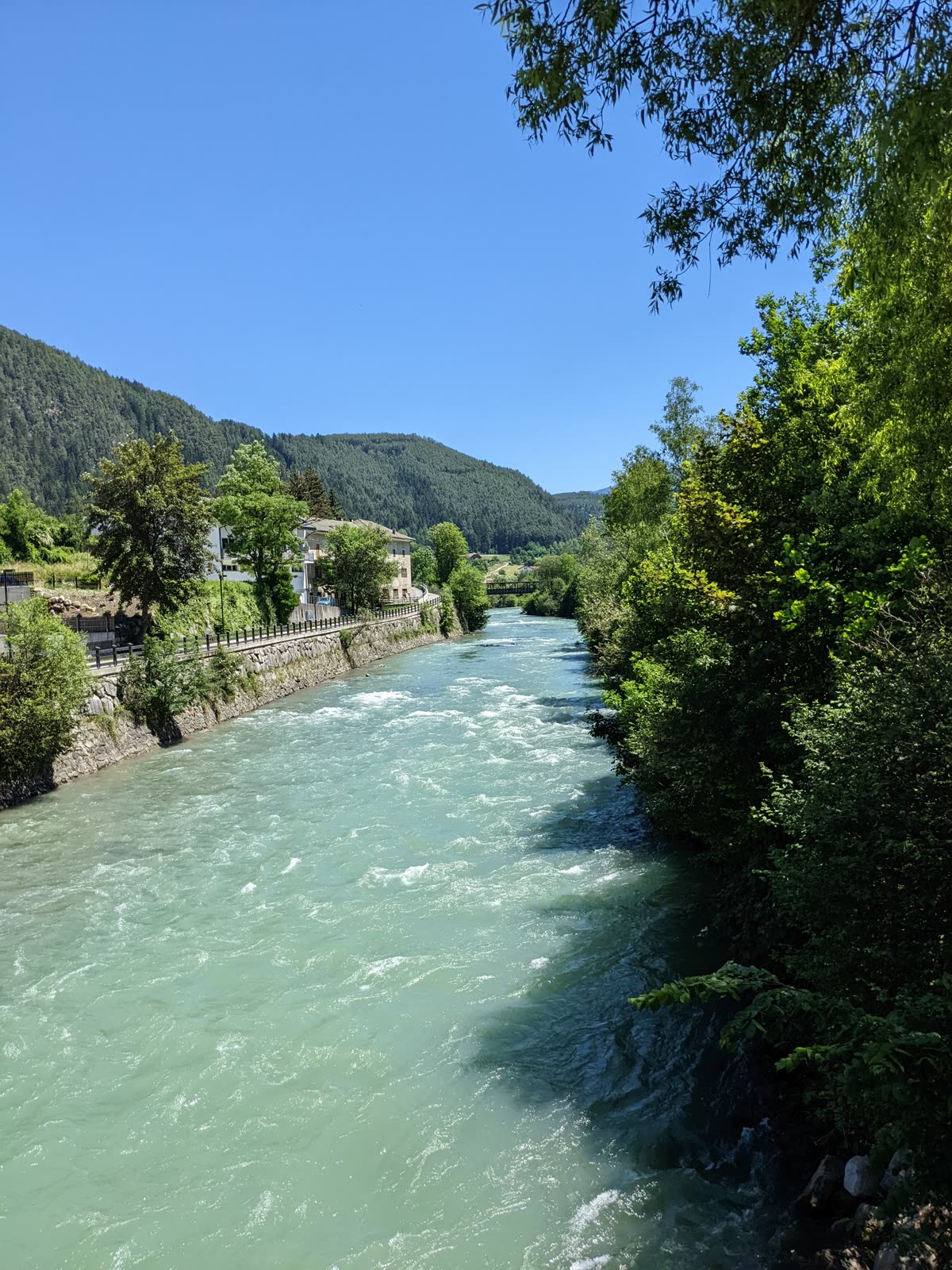 Fast turquoise alpine river running between wooded banks, houses and a footbridge upstream, forested ridges beyond