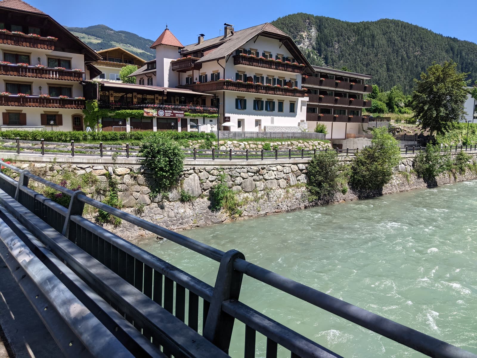 View from a bridge toward South Tyrolean guesthouses with flower-filled balconies above the turquoise river, a Bikes Welcome sign on the terrace