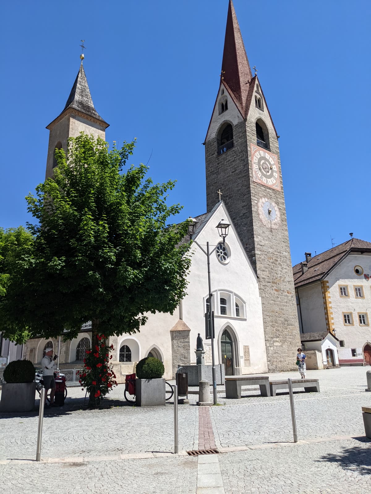 Parish church of San Lorenzo di Sebato with two mismatched bell towers, a white Gothic gable with rose window, and a painted clock face, on a cobbled square