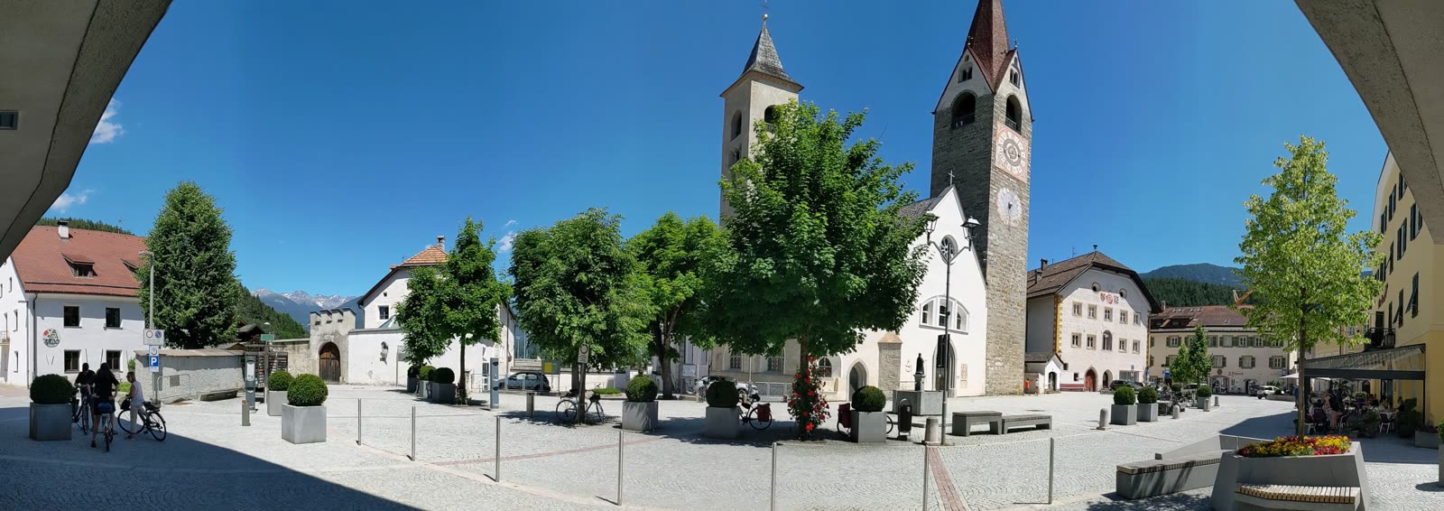 Panorama of the cobbled main square of San Lorenzo di Sebato with the twin-towered church, frescoed houses, trees, benches and loaded touring bikes