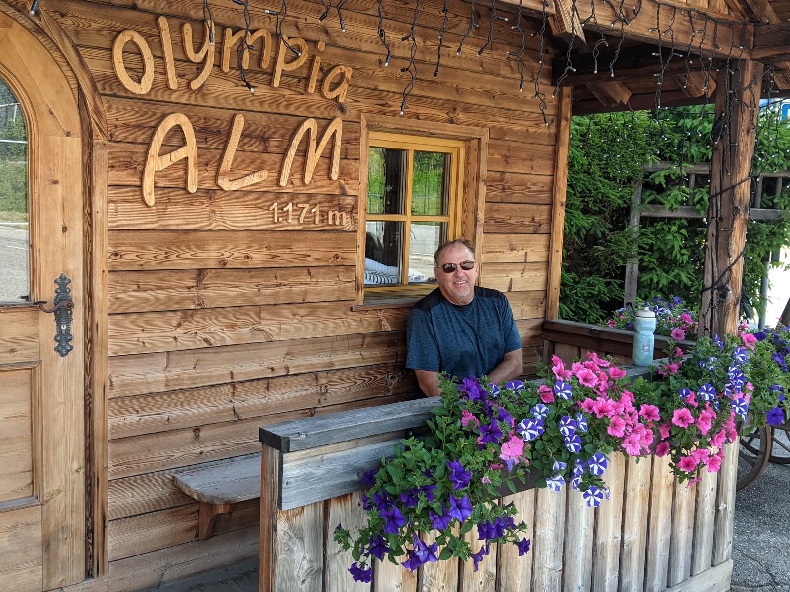Mike sitting at a wooden alpine hut with OLYMPIA ALM 1.171 m carved into the timber wall, flower boxes of pink and purple petunias in front