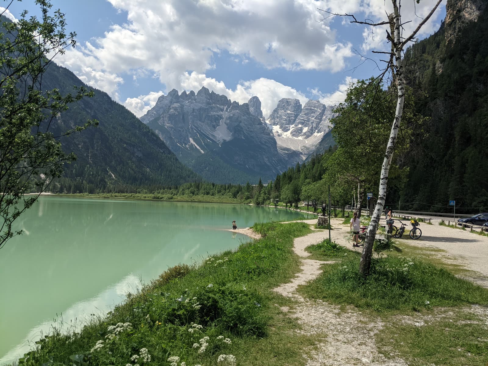 Glacier-green calm alpine lake reflecting the jagged Monte Cristallo massif, a gravel path and touring bikes along the near shore