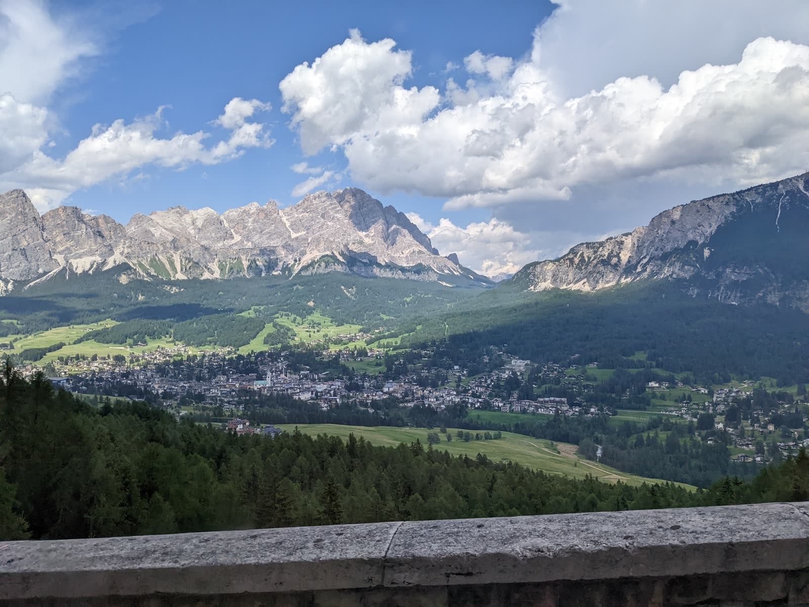 Elevated view over Cortina d'Ampezzo spread across its green valley bowl, the town campanile at center, ringed by pale Dolomite peaks under building clouds