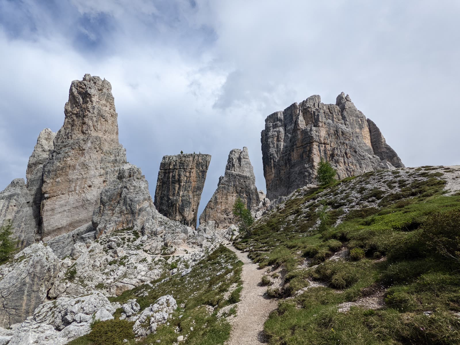 The full cluster of the Cinque Torri rock towers rising from a grassy shoulder, a footpath leading up to their base under a moody grey sky