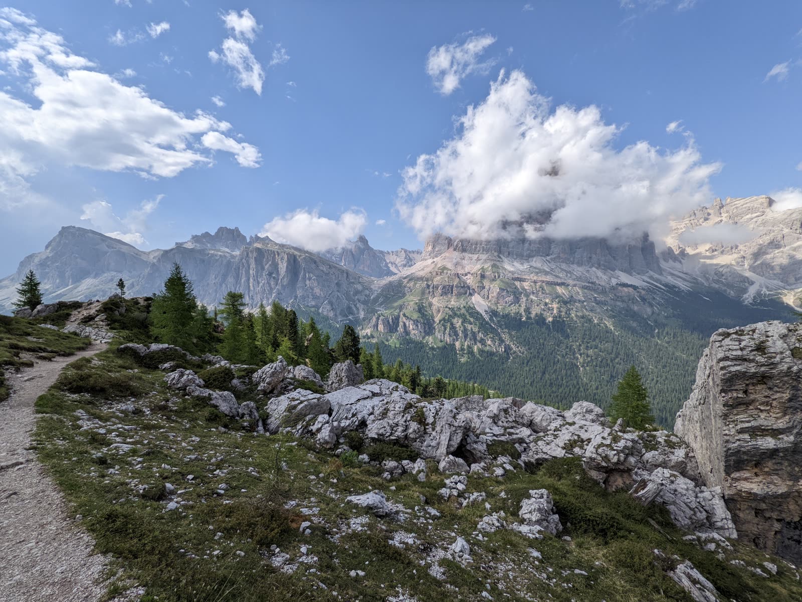 View out from the Cinque Torri shoulder over larches and white rock toward ridge after ridge of hazy Dolomite peaks under a big cloud