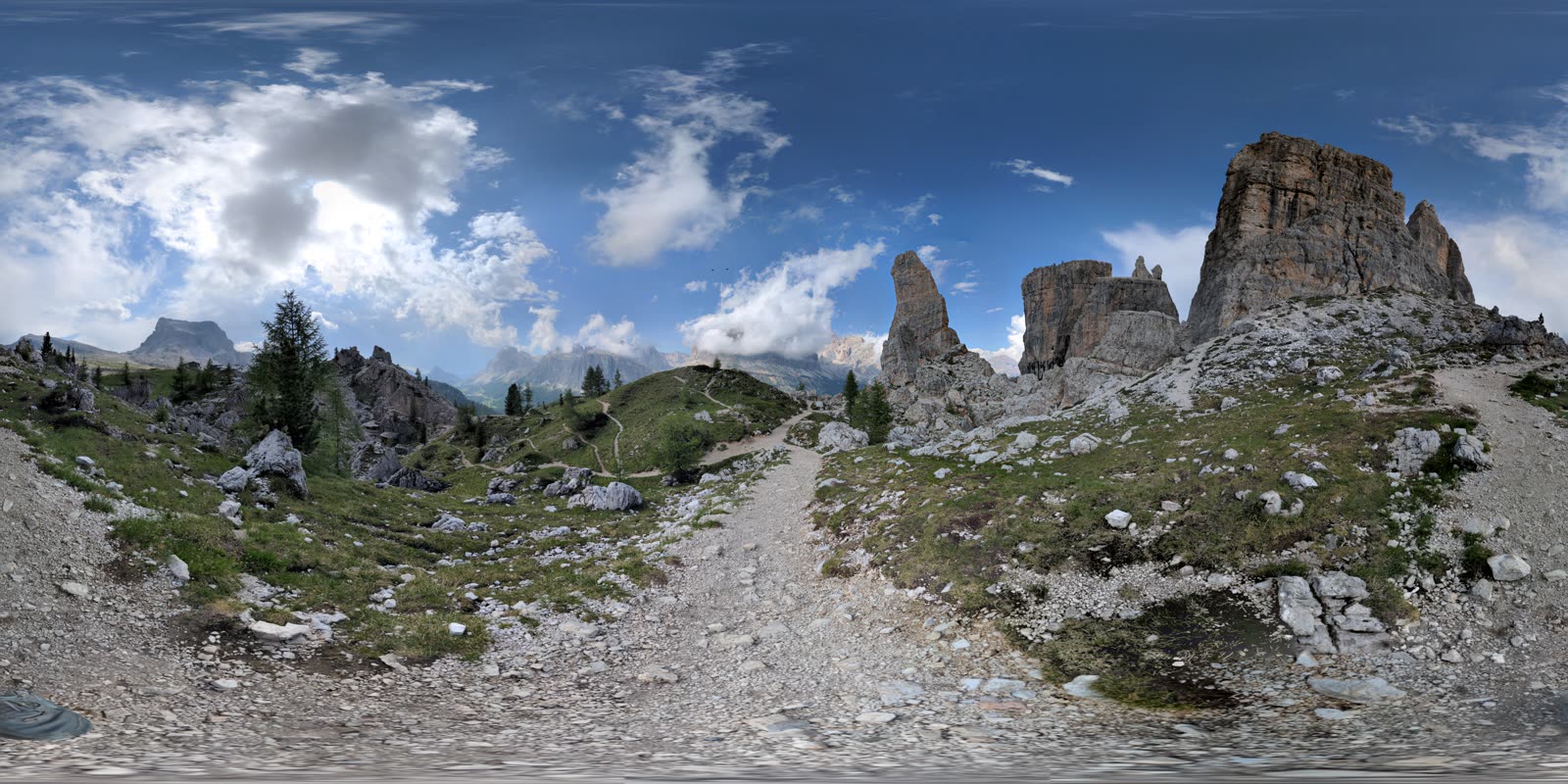 A 360-degree spherical panorama of the Cinque Torri, the rock towers curving up on the right, footpaths and meadow bending around the whole frame under a wide blue sky