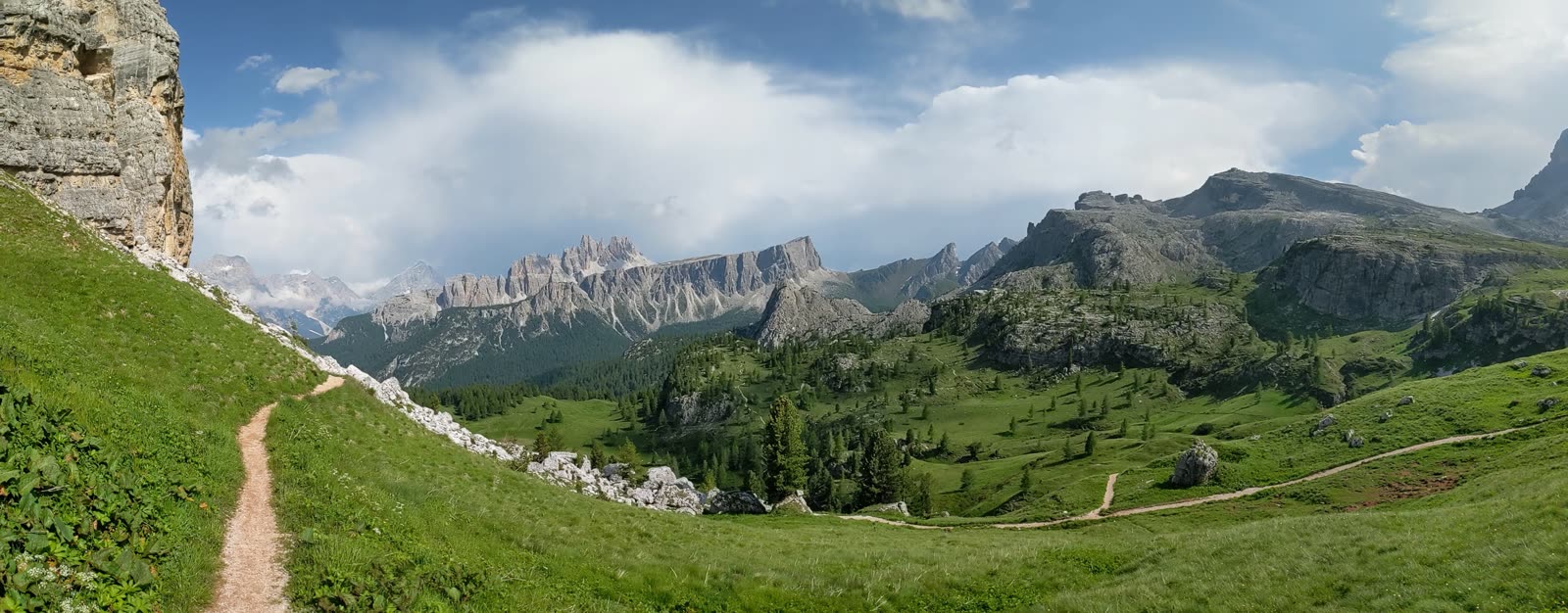 Wide panorama of the Cinque Torri high meadow, a footpath curving through green grass toward a horizon ringed by jagged grey Dolomite peaks and ridges