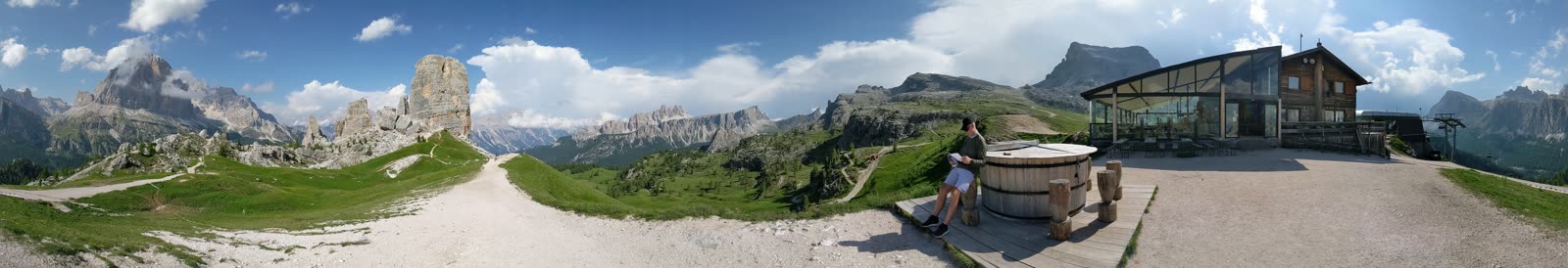 Ultra-wide panorama from the top of the Cinque Torri gondola: the rock towers at left, open meadow and Dolomite ridges across the middle, and the glass-and-timber Rifugio Scoiattoli with its terrace at right
