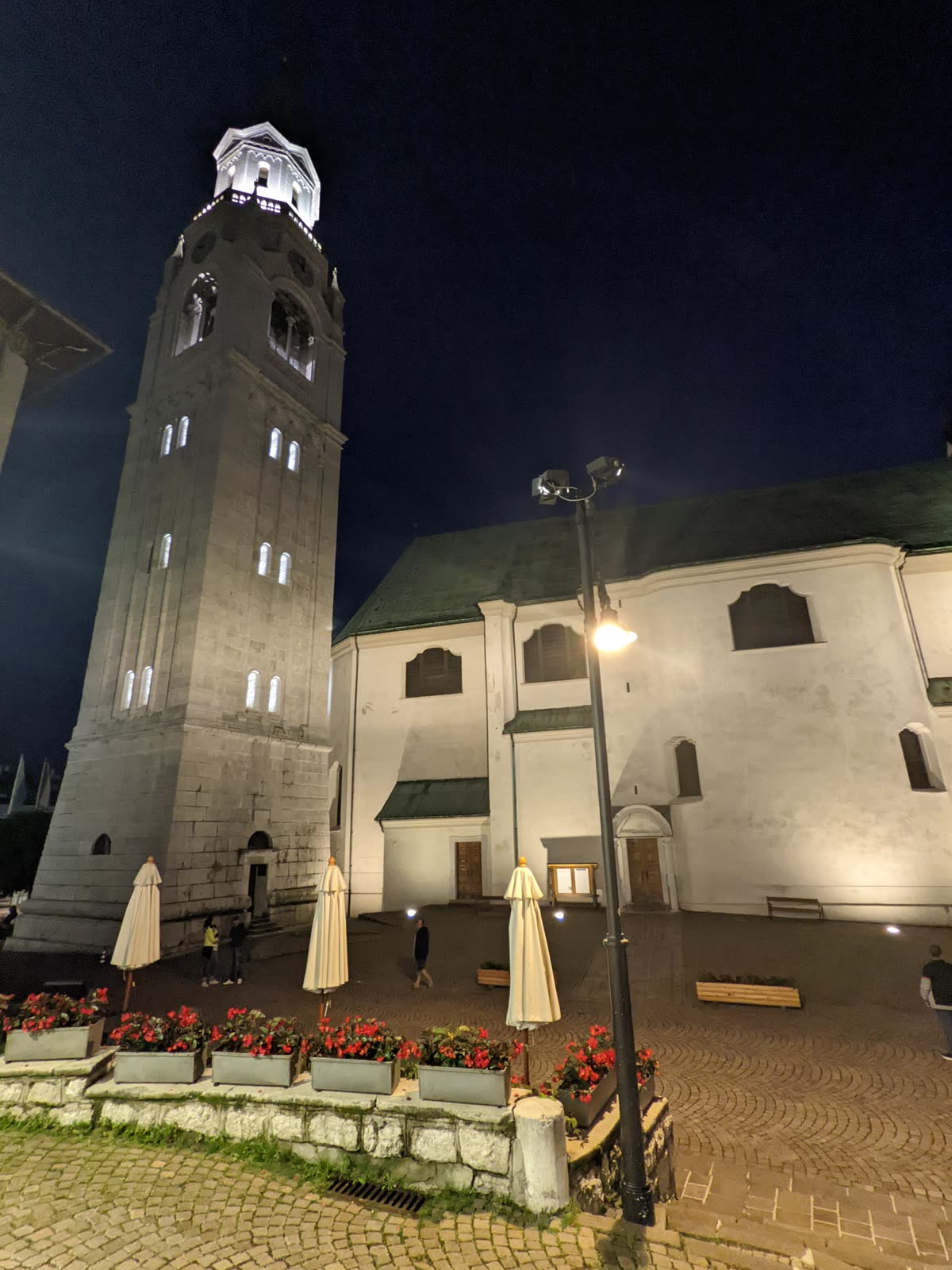 Cortina's tall stone bell tower floodlit white against a deep blue night sky, the parish church beside it, closed café umbrellas and red flower boxes in the cobbled square below