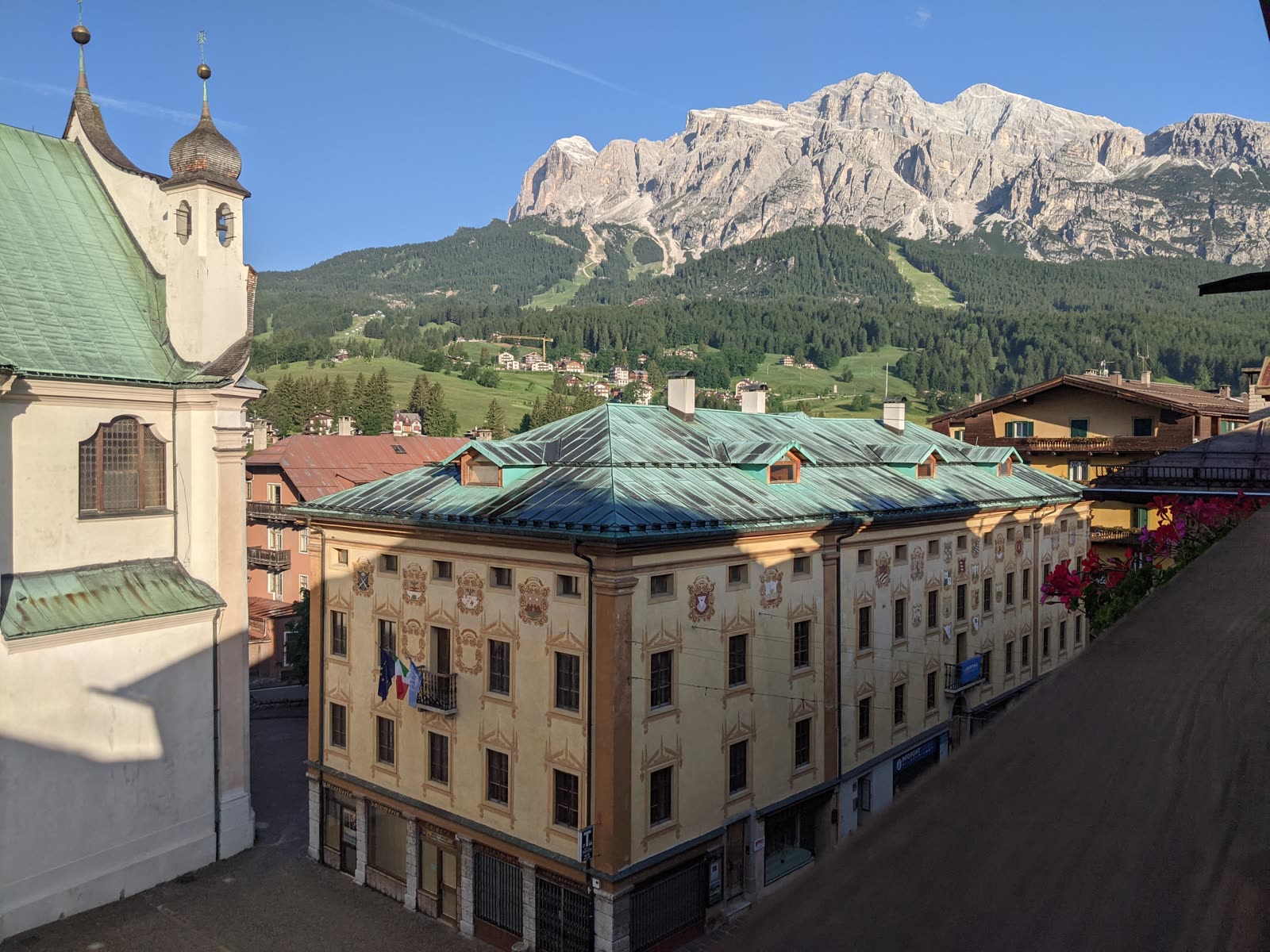Early-morning view from a Cortina window over green copper rooftops and a church tower to the huge pale wall of the Tofana massif catching first sun under a clear blue sky
