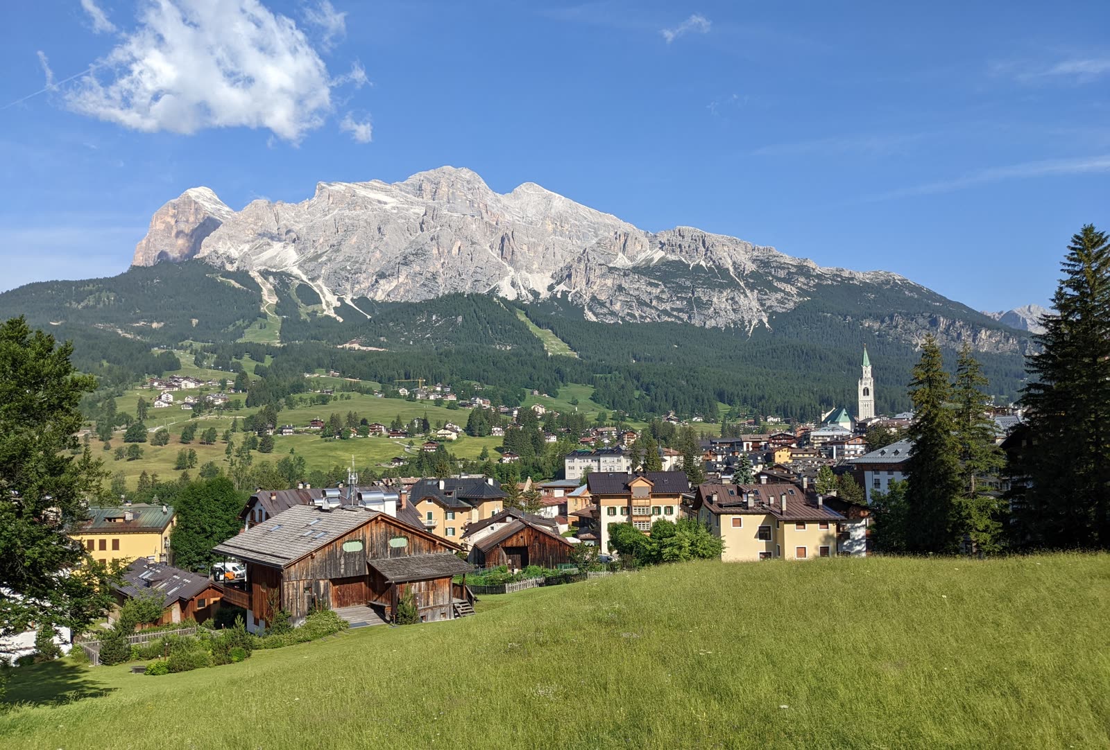 Cortina spread across its green valley bowl with its church campanile at right, wooden chalets and meadows in the foreground, the pale Tofana massif rising behind under blue sky