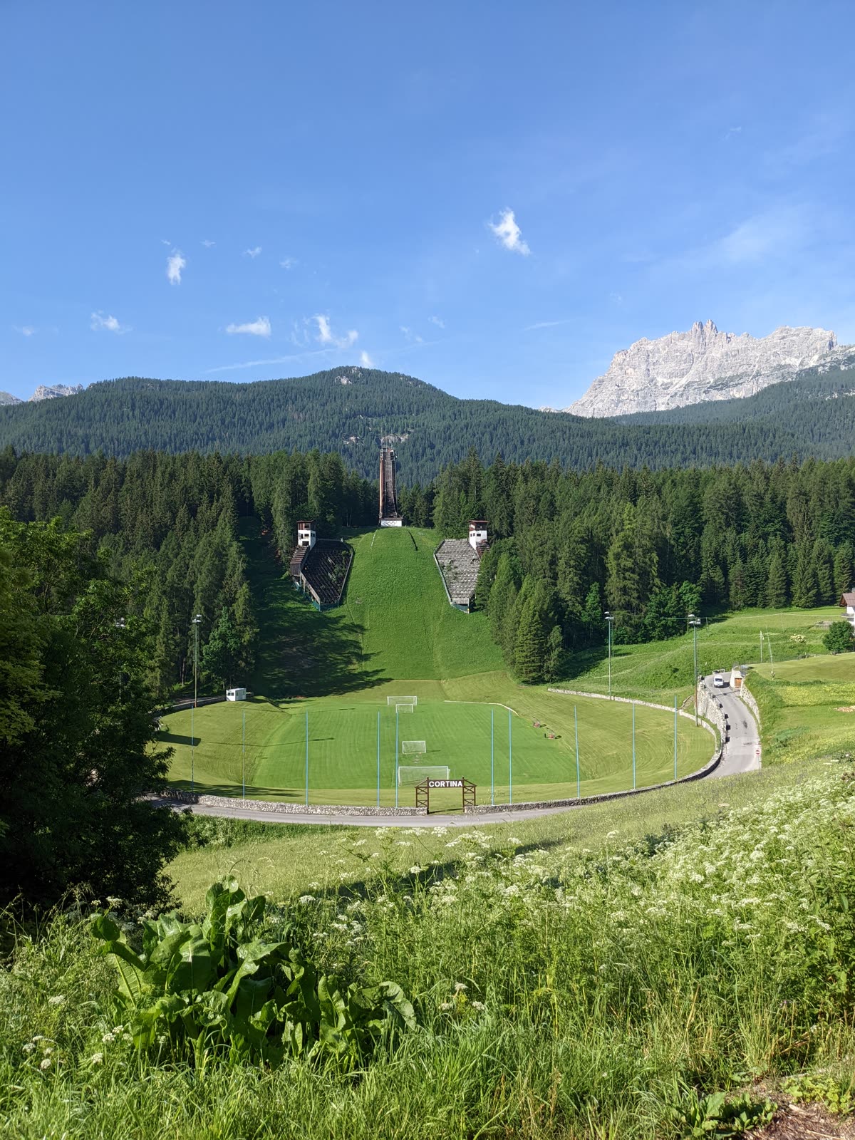 A tall abandoned Olympic ski-jump inrun tower rising from a steep green grassed-over landing hill, old timber judges' stands on either side, forested Dolomite ridges behind