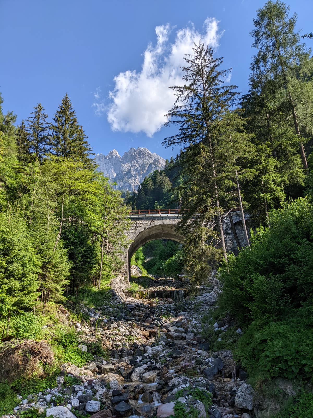 An old stone railway arch bridge spanning a dry boulder-strewn streambed, tall spruce and larch on both banks, jagged grey Dolomite peaks rising in the distance under blue sky and a white cloud