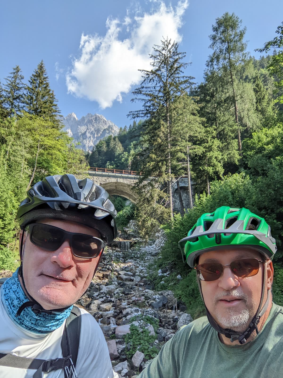 Selfie of Mike and Doug in cycling helmets and sunglasses in front of the stone railway arch bridge, forest and a distant grey Dolomite peak behind them