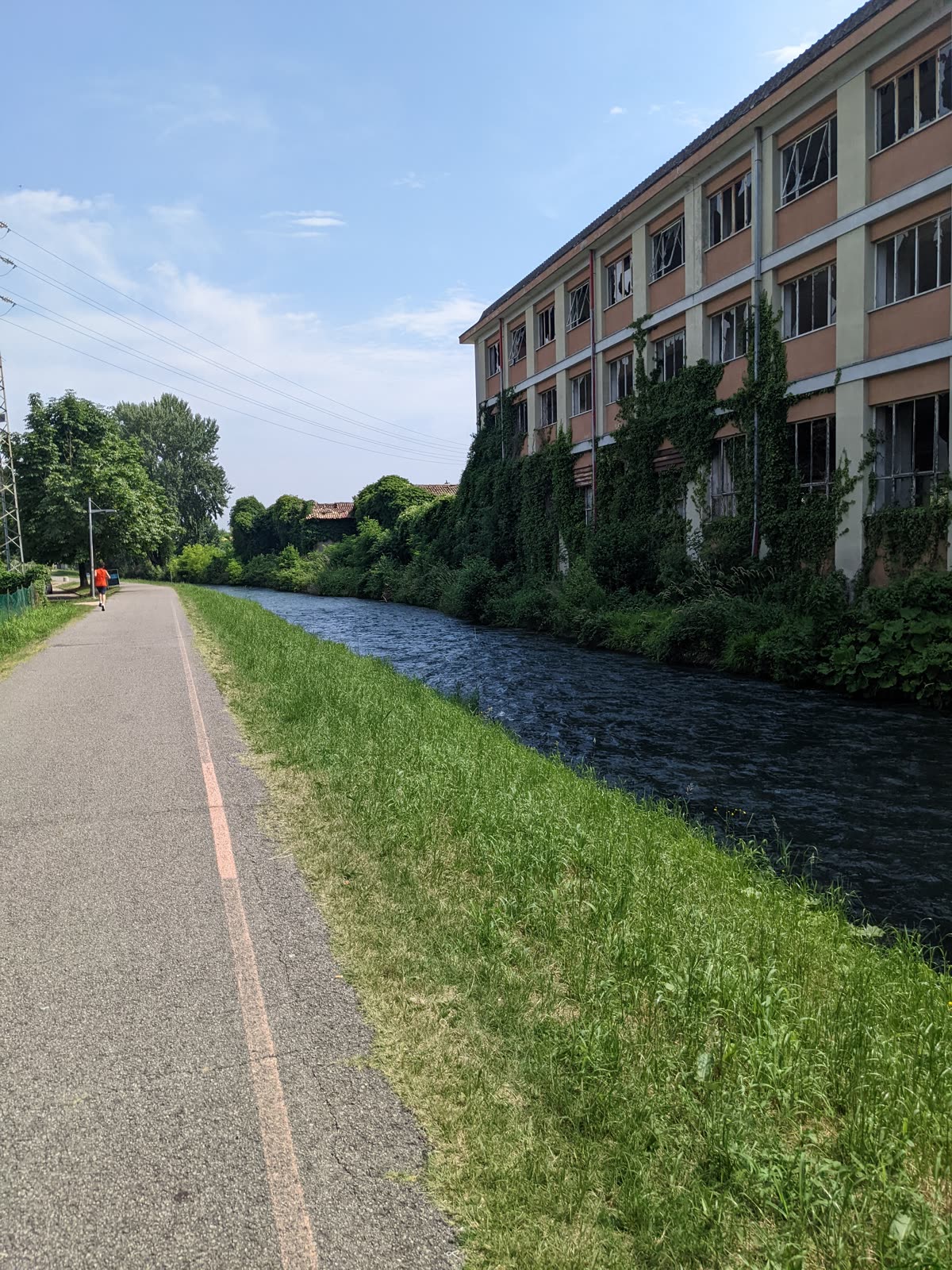 A flat paved cycle path running dead straight beside a slow dark canal at Vittorio Veneto, a jogger ahead in orange, a long ivy-covered abandoned factory across the water under a hazy warm sky
